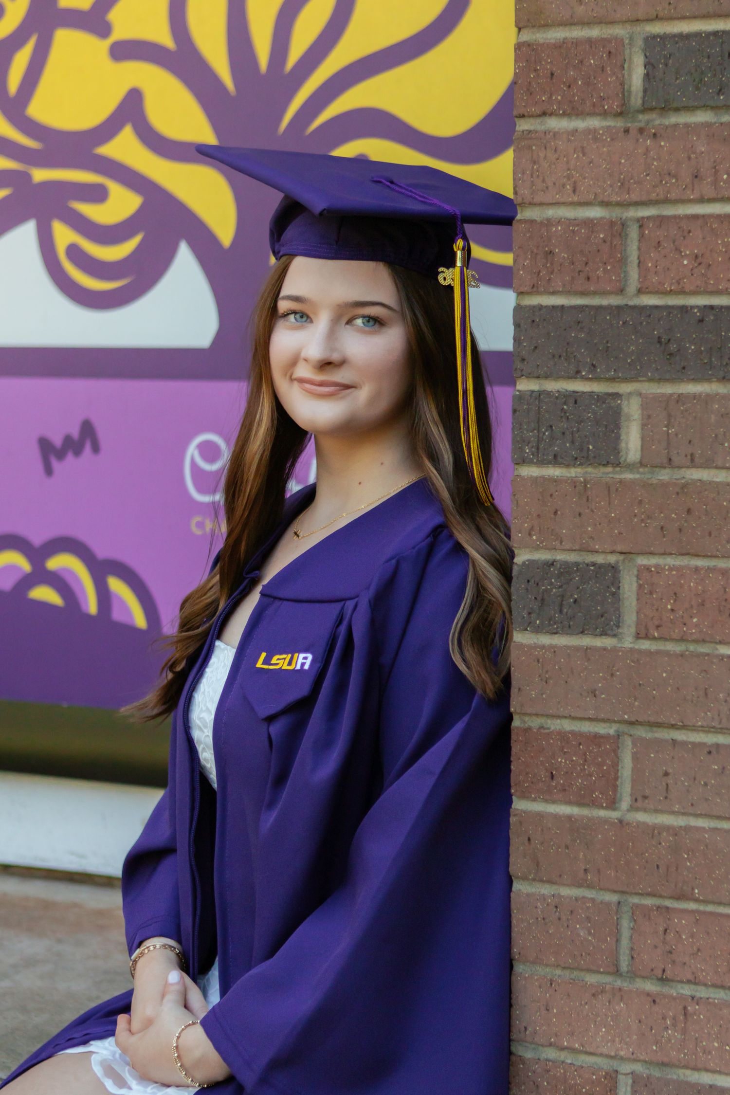 Female student in purple LSU graduation cap and gown leaning against brick wall for senior portrait.
