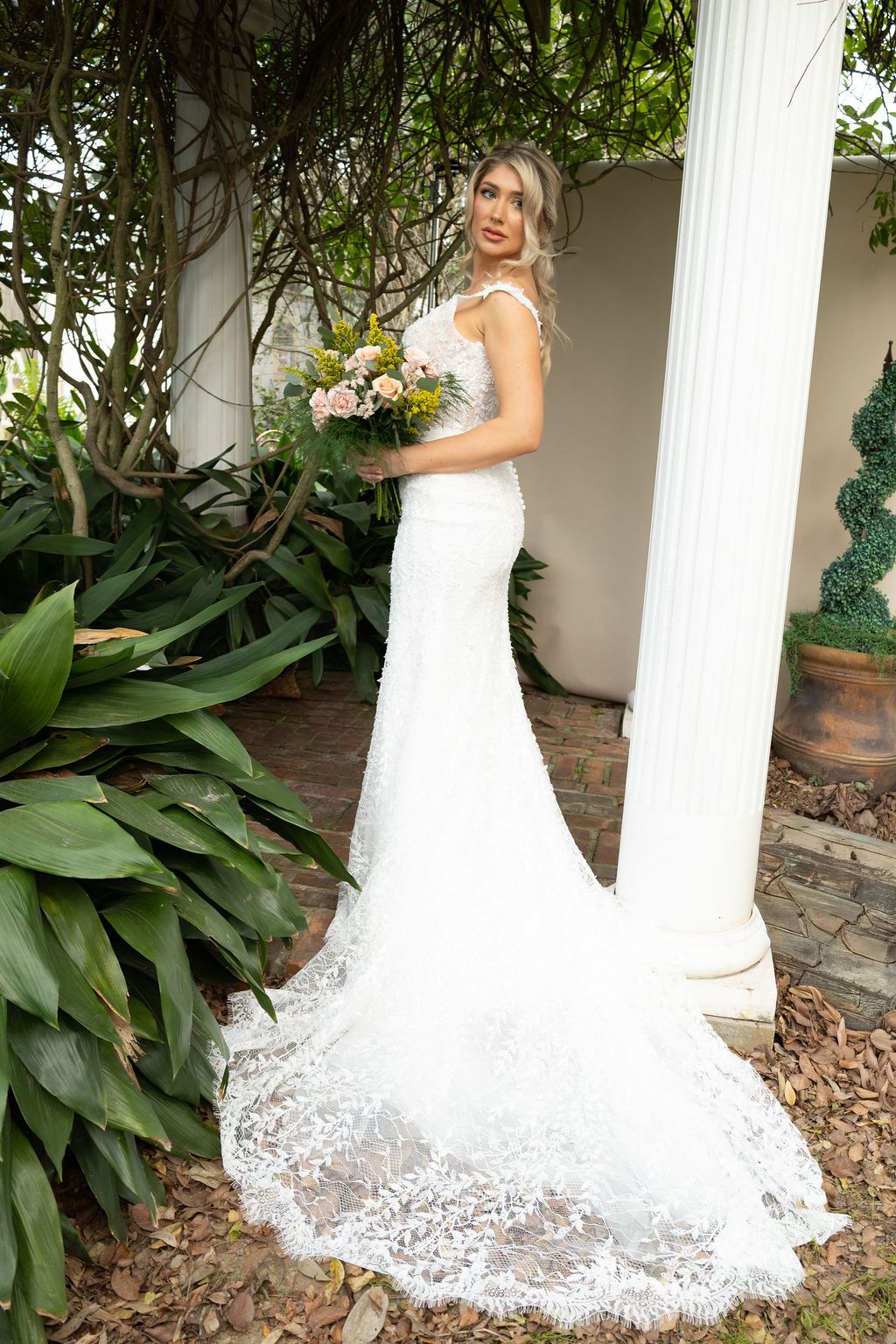 Bride in white lace mermaid wedding gown with train holds bouquet beside garden column.