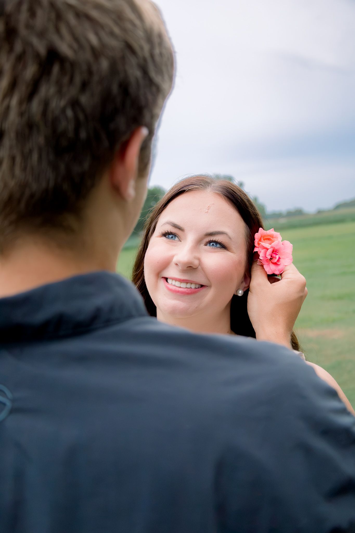 Man placing a pink flower behind a smiling woman's ear in a green field setting.