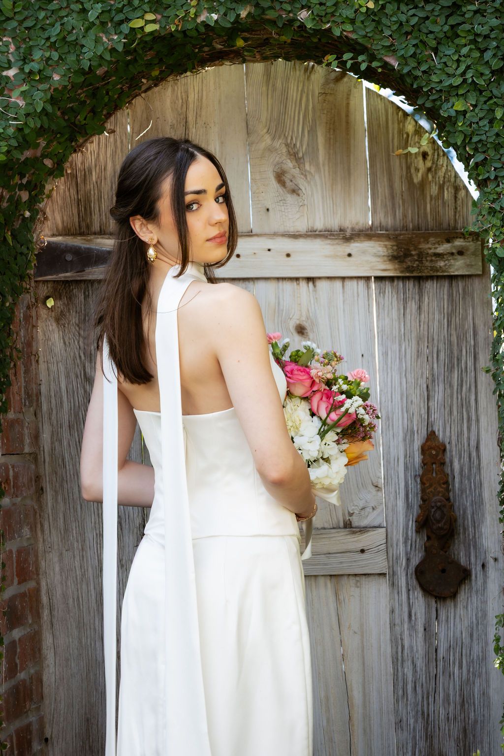 Bride in white halter gown holding a pink bouquet, posing at a rustic wooden arched door covered in ivy.