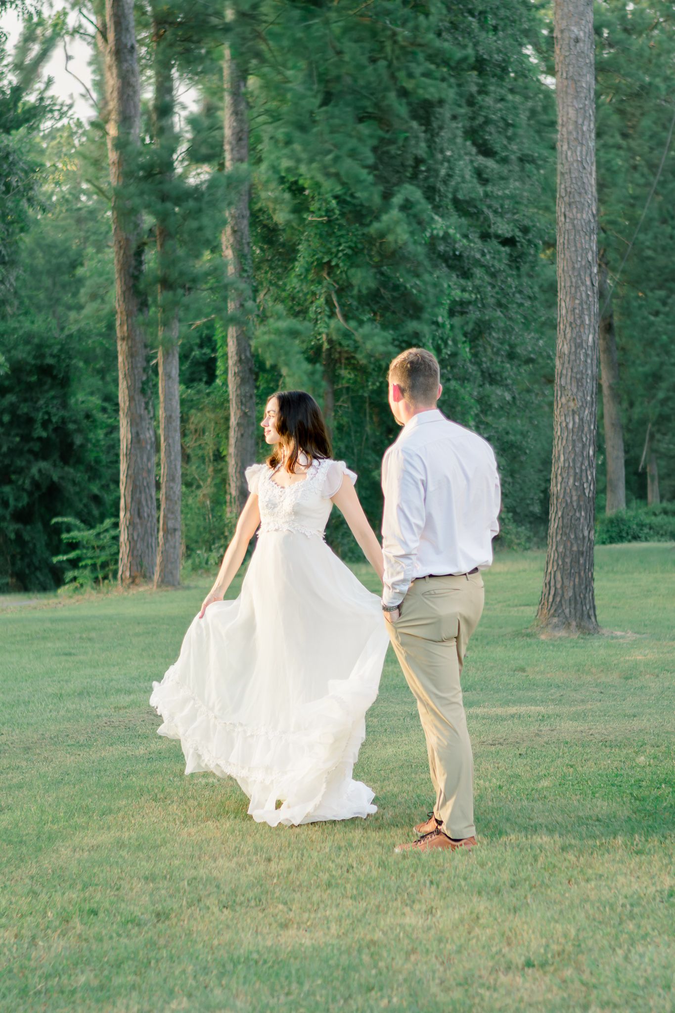 A bride in a flowing white dress and groom in khaki walk hand in hand across a lush green lawn surrounded by tall trees.