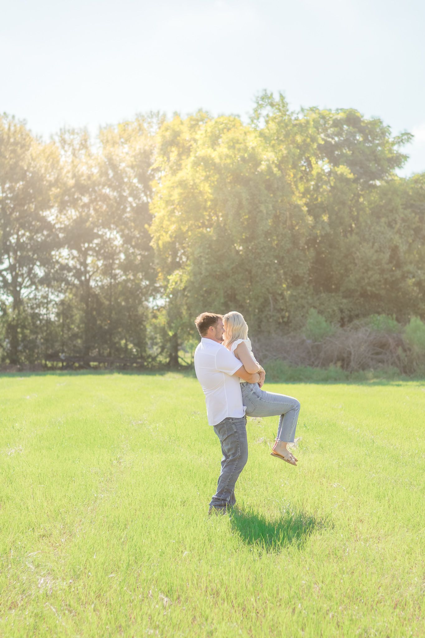 A couple shares a romantic kiss in a sunlit green field, with the man lifting the woman off the ground near lush trees.