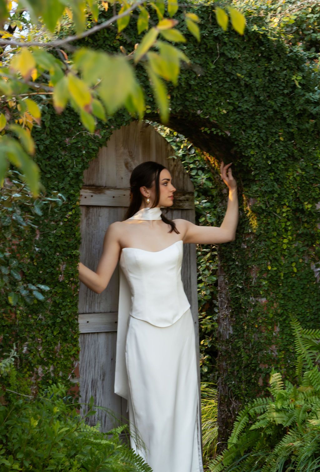 Elegant bride in strapless white gown standing in an ivy-covered stone archway with a wooden door.