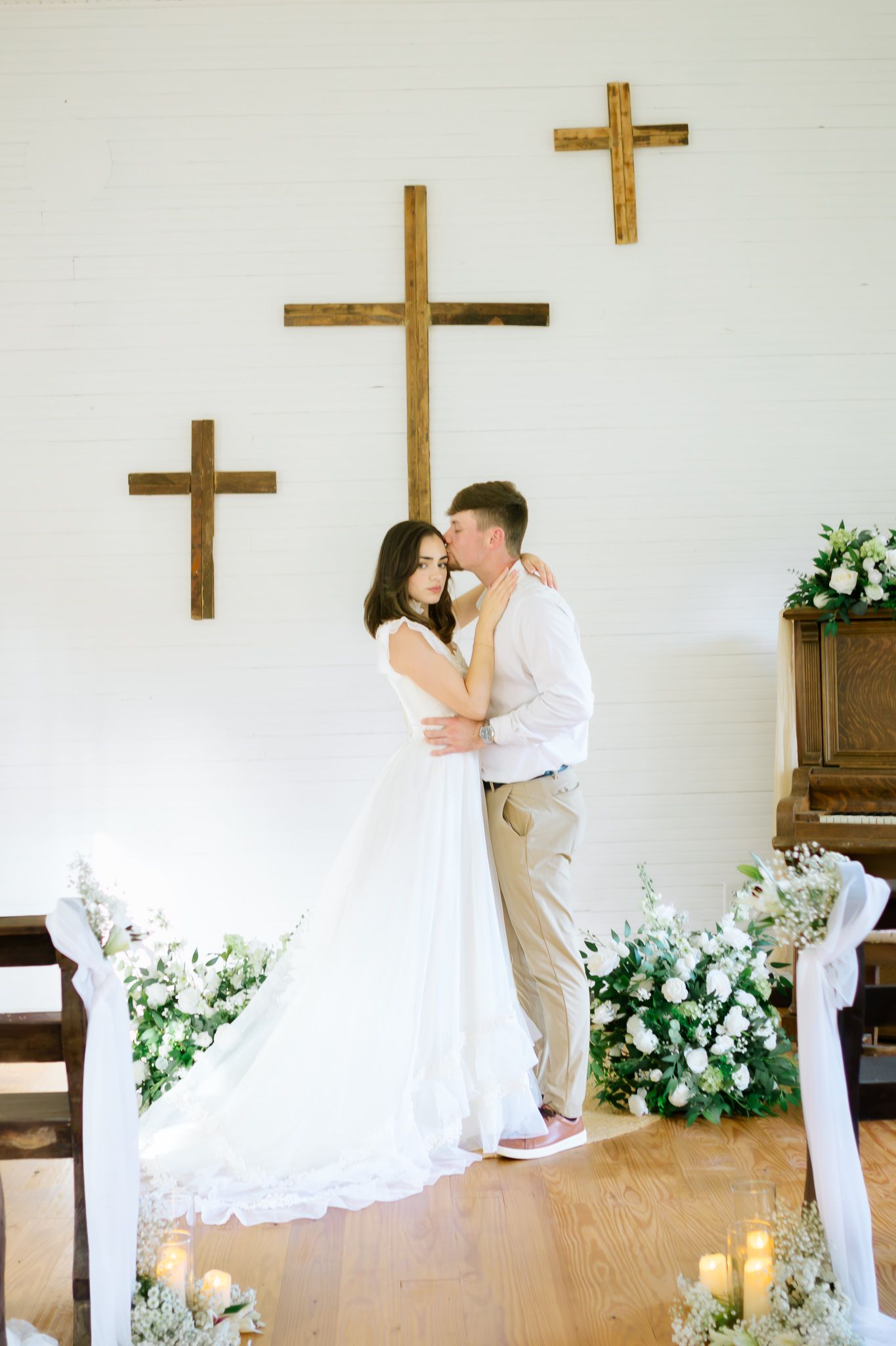 Bride and groom embracing at church altar with wooden crosses, white floral arrangements, and candles.