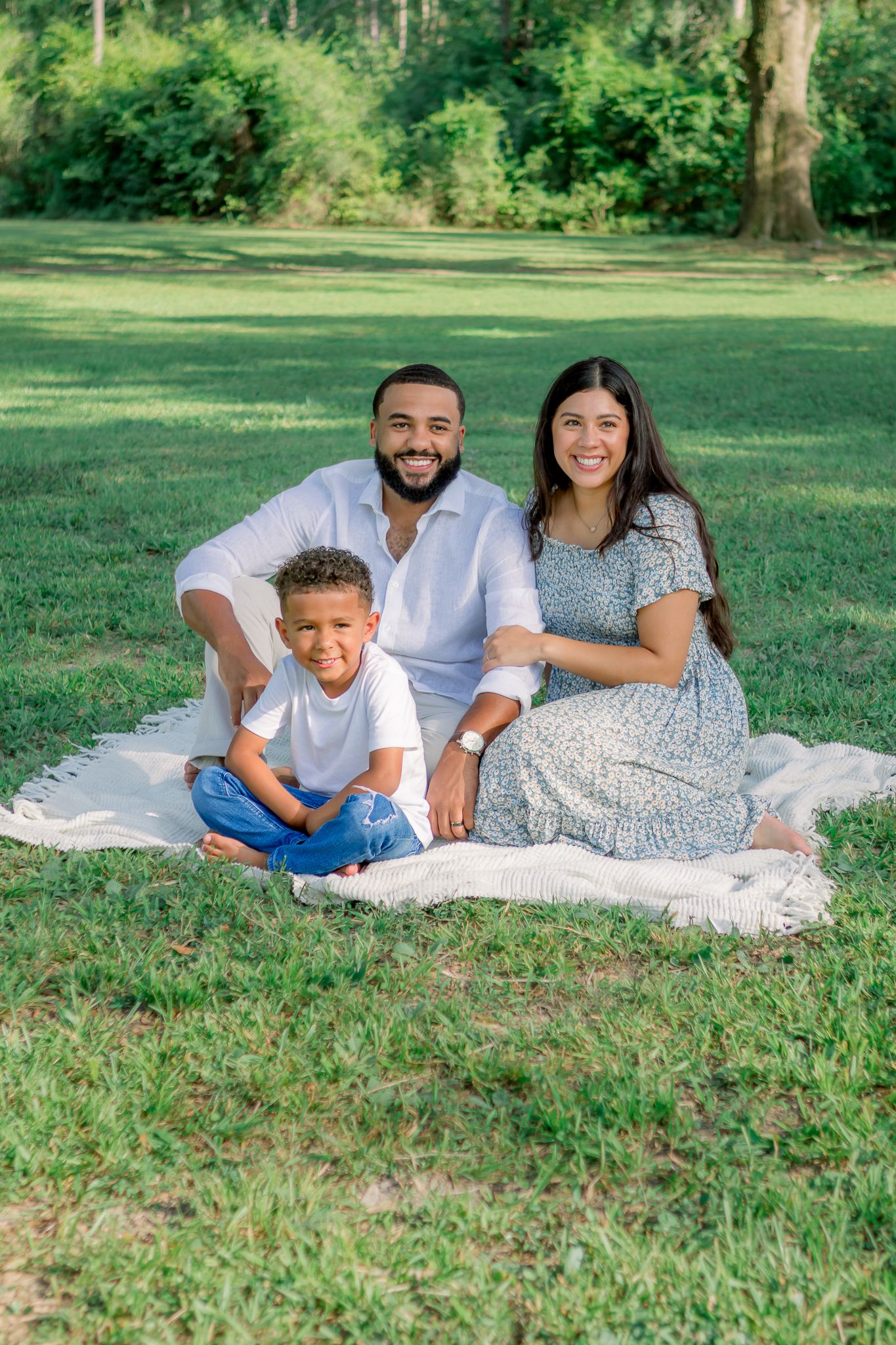 A happy family of three sits on a blanket in a green park, smiling warmly for a portrait photo.