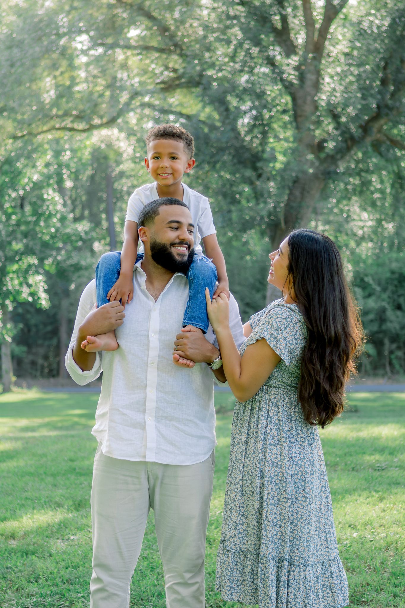 A happy family poses outdoors, father carrying son on shoulders while mother smiles, surrounded by lush green trees.