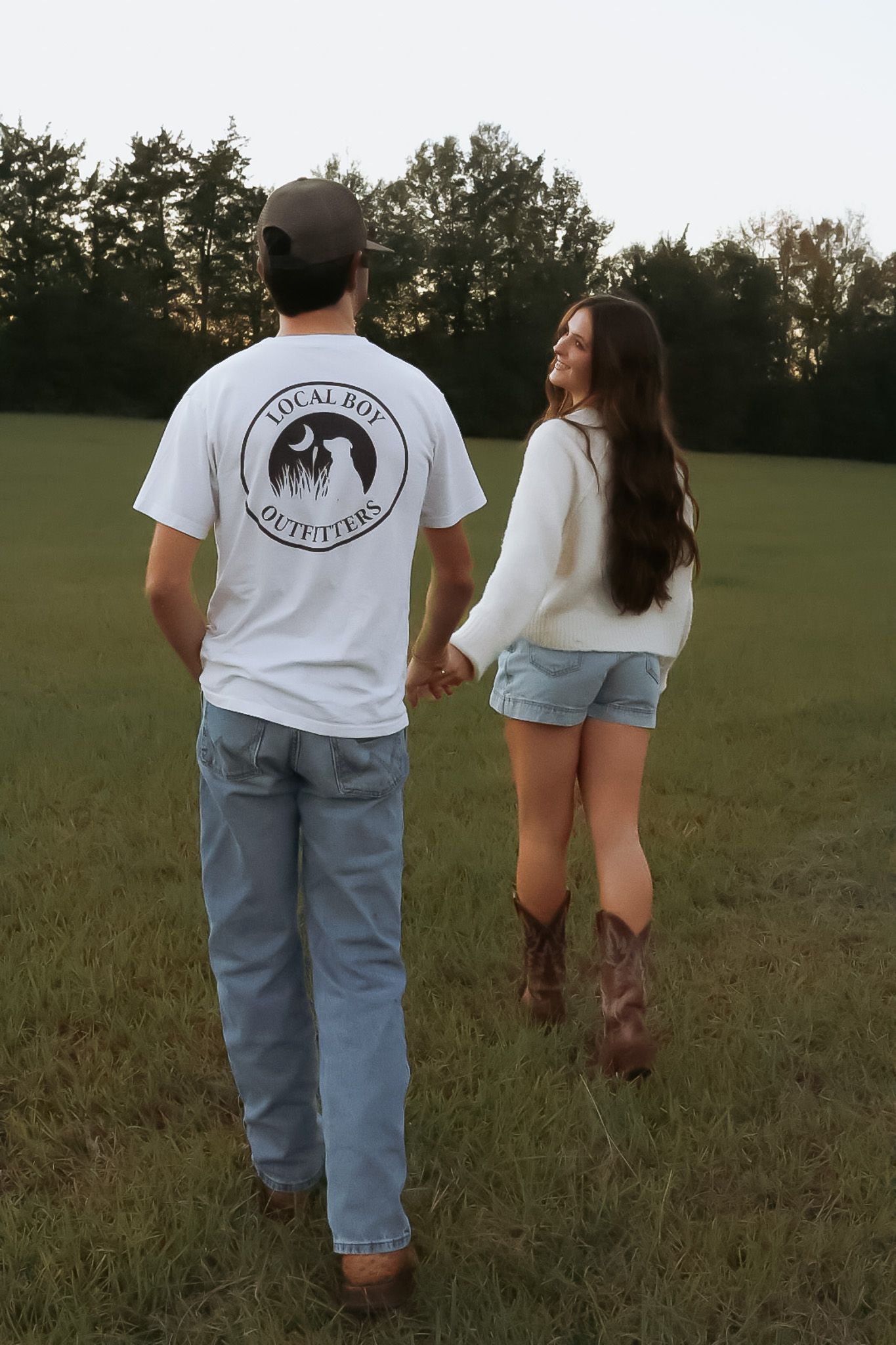 Couple walking hand in hand through a field wearing matching white t-shirts and jeans.