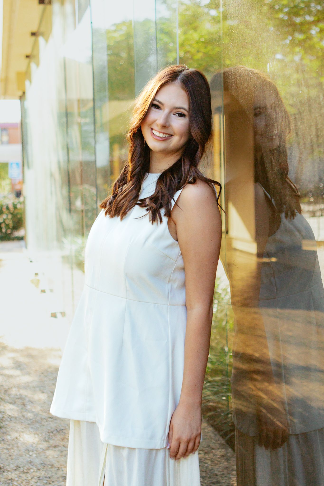 Smiling young woman in white sleeveless dress leaning against a glass wall with her reflection visible.