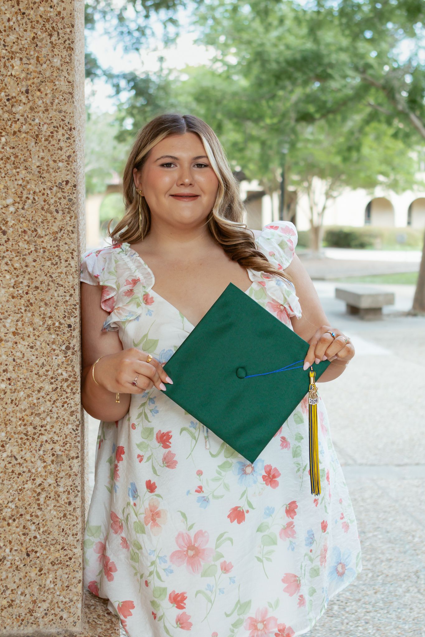 Young woman in floral dress holding a green graduation cap while leaning against a stone column outdoors.