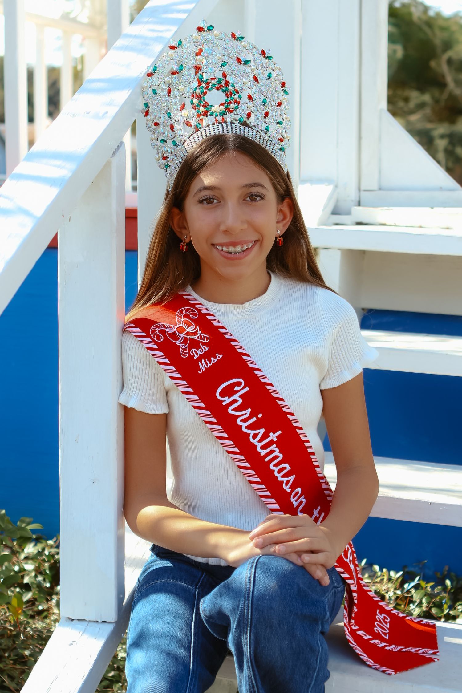 Person wearing white shirt and jeans sits on white porch railing with red sash.