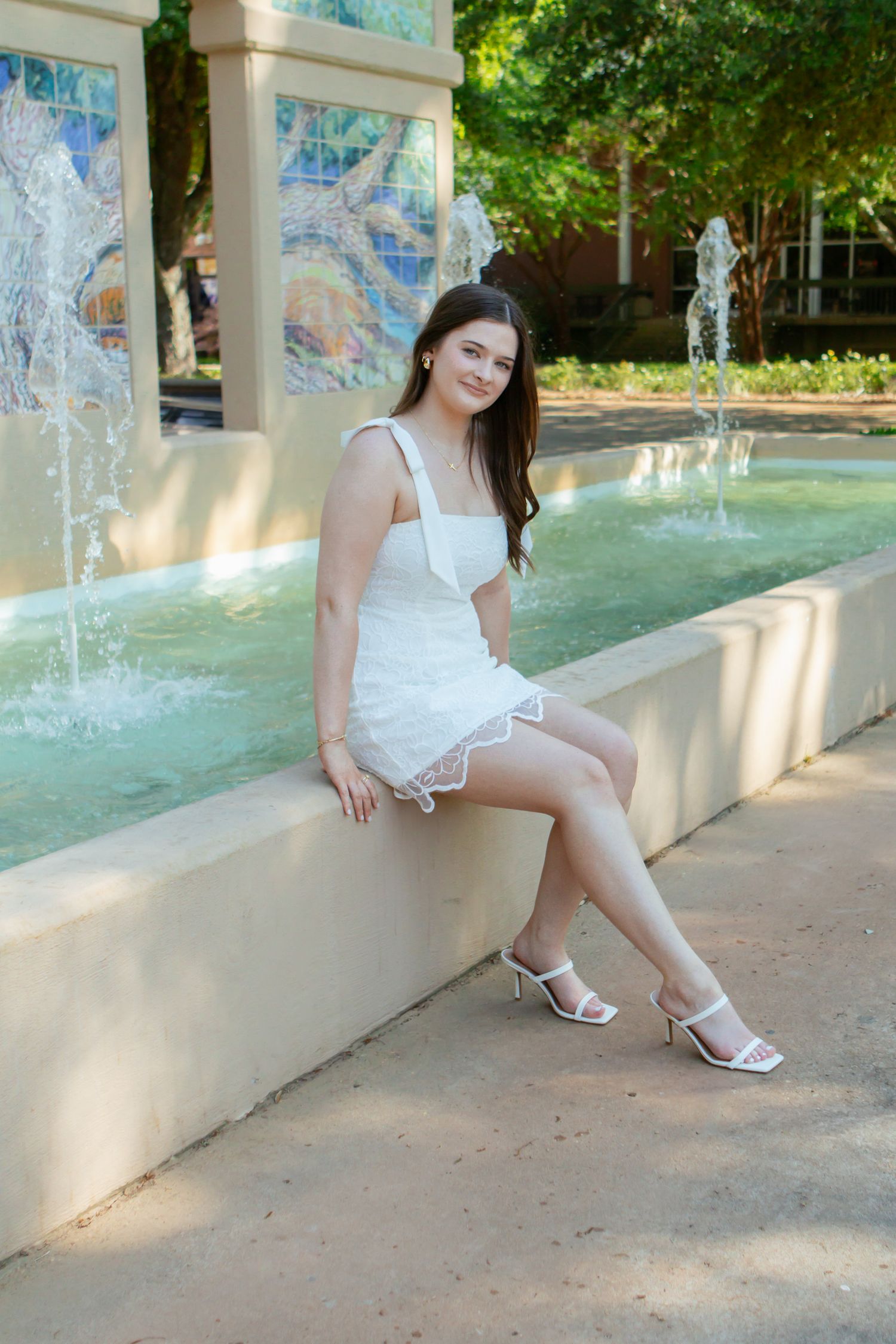 Young woman in white mini dress sitting by decorative fountain with mosaic tile backdrop on campus.