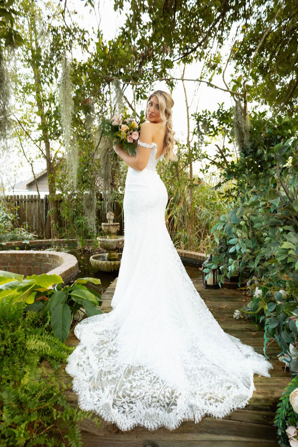 Elegant bride in backless lace mermaid gown with long train poses in lush garden setting.