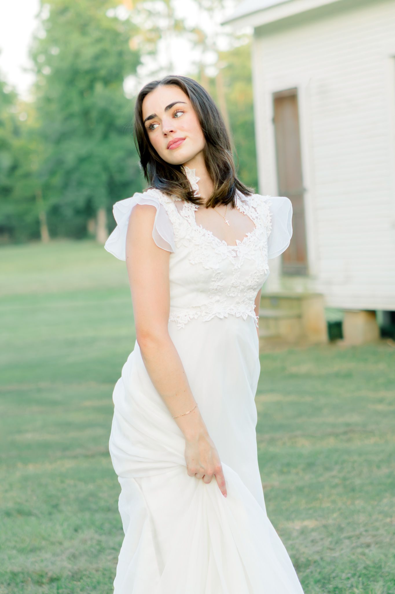 A bride in a white lace wedding gown with flutter sleeves poses outdoors on green grass near a white building.