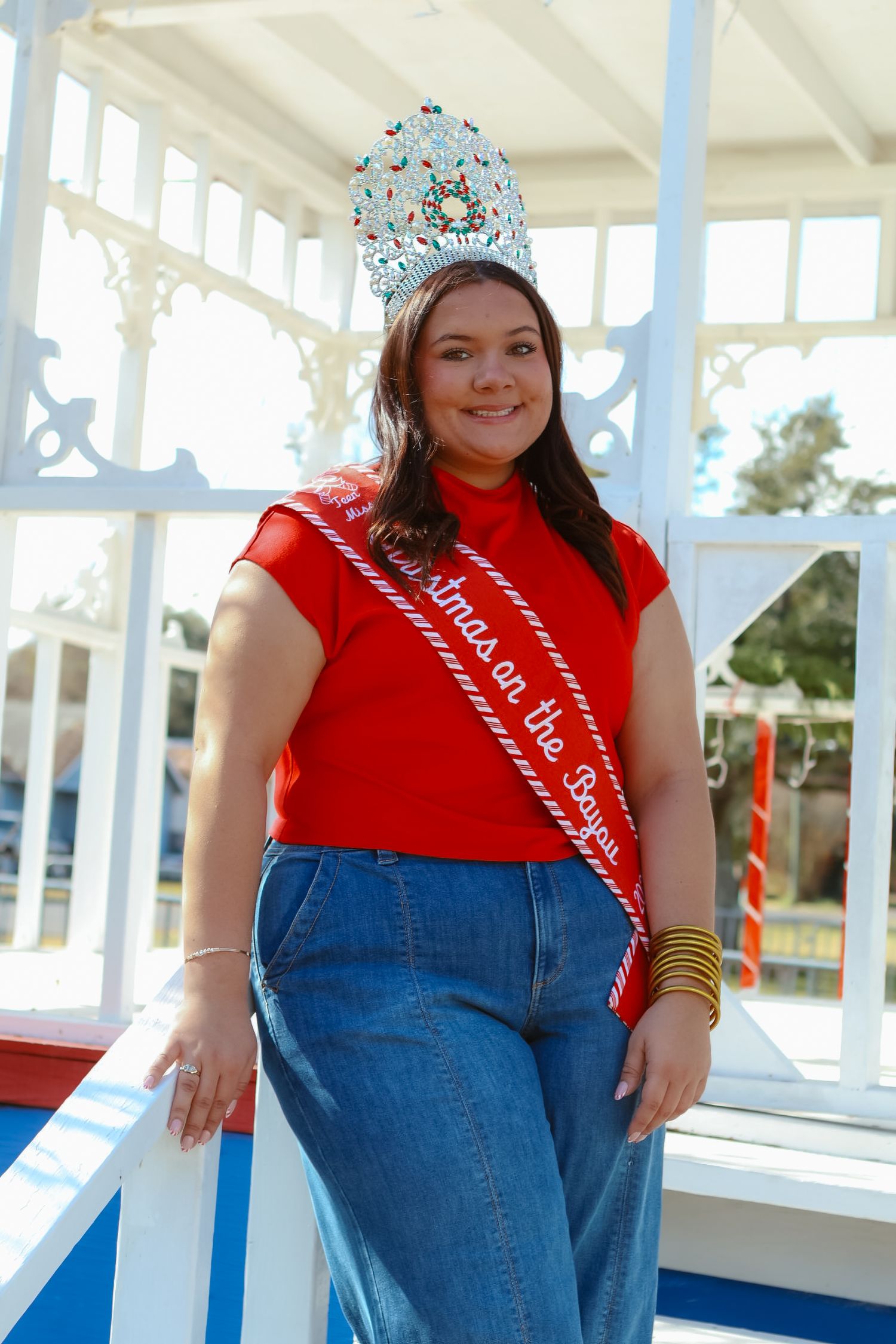 Person in red shirt and jeans poses on white porch with decorative crown.
