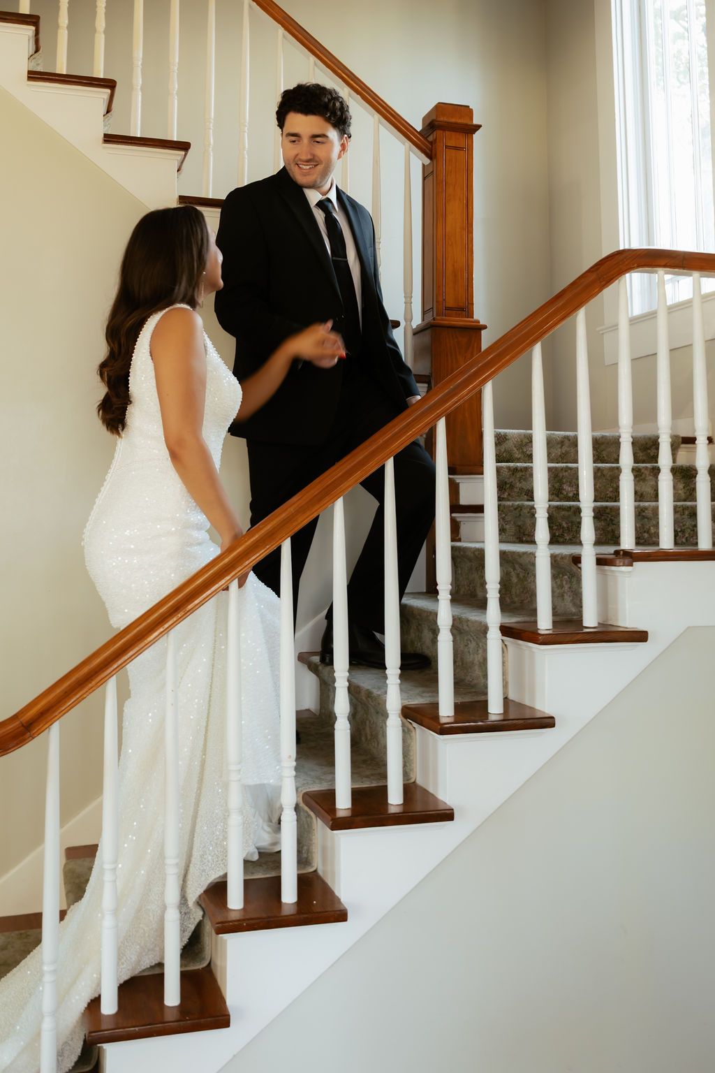 Smiling groom in black suit greets bride in white gown on elegant staircase.