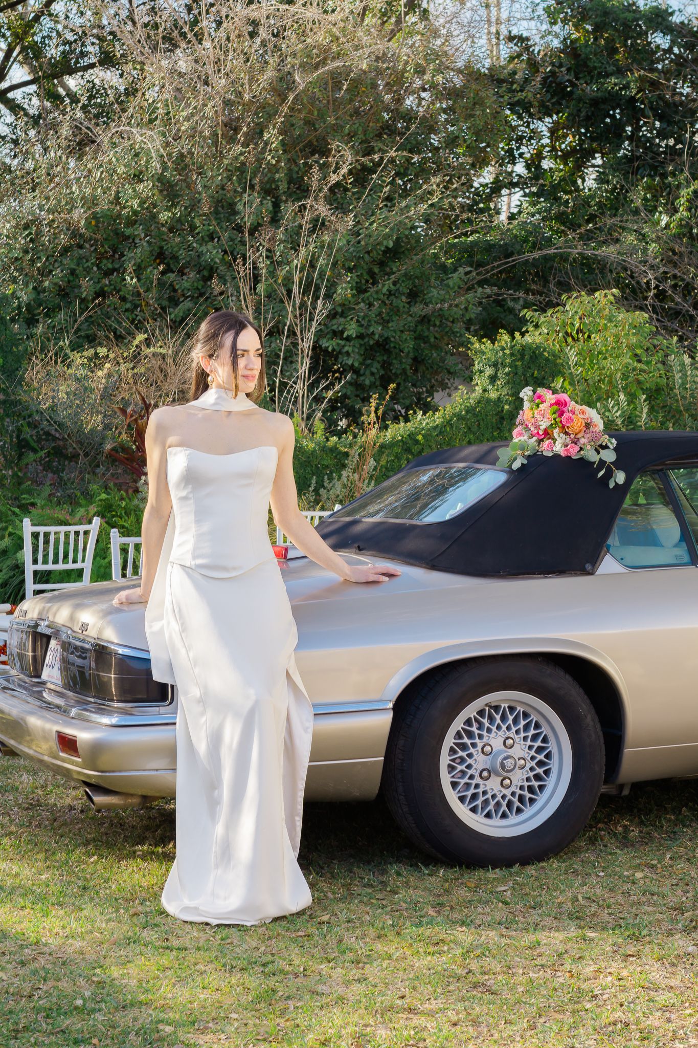 Elegant bride in a white halter gown leaning against a vintage silver car adorned with a colorful floral bouquet.