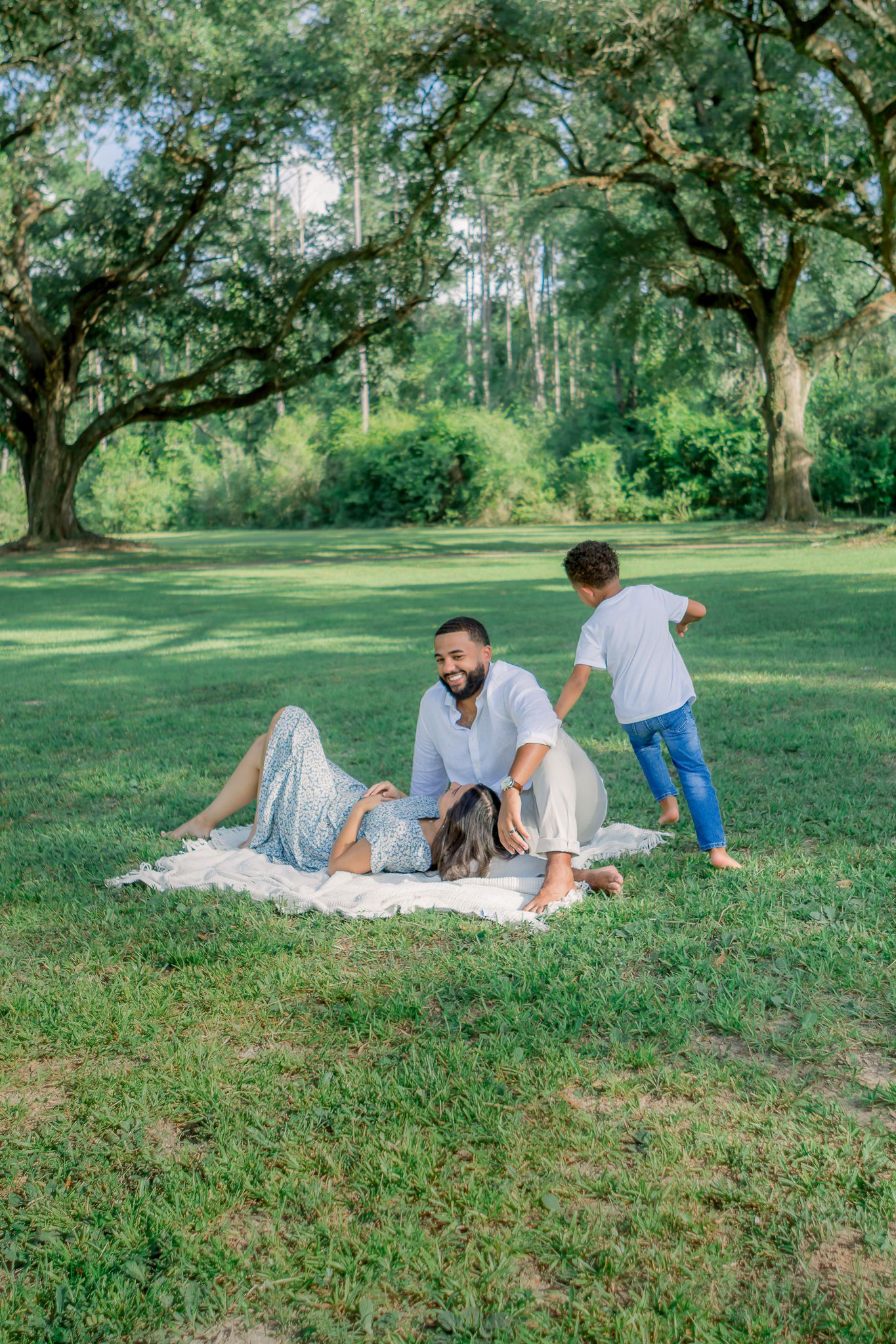 Family enjoying a sunny day picnic on a blanket in a lush green park with large oak trees.