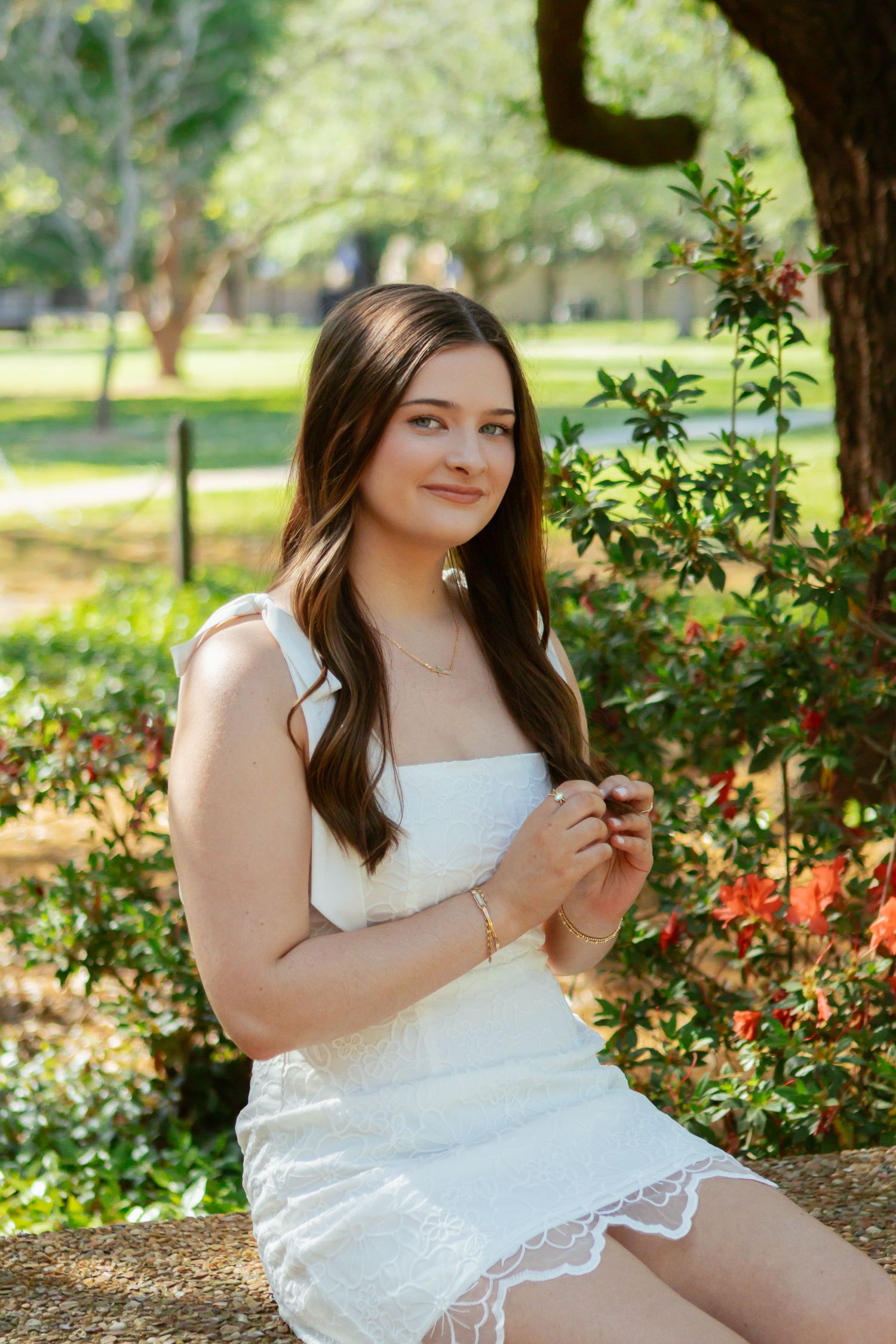 Young woman in white lace mini dress sitting on stone wall surrounded by red flowering bushes outdoors.