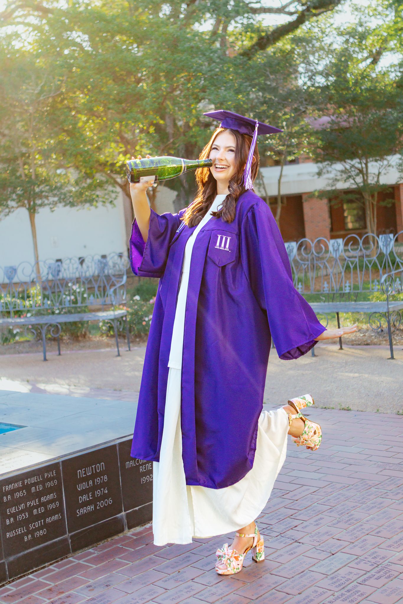 Graduate in purple cap and gown celebrating outdoors holding a bottle on a sunny campus plaza.