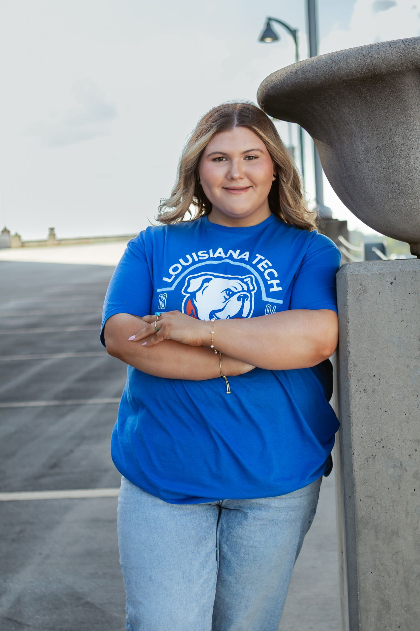 Young woman in Louisiana Tech blue t-shirt and jeans leaning against a concrete pillar with arms crossed.