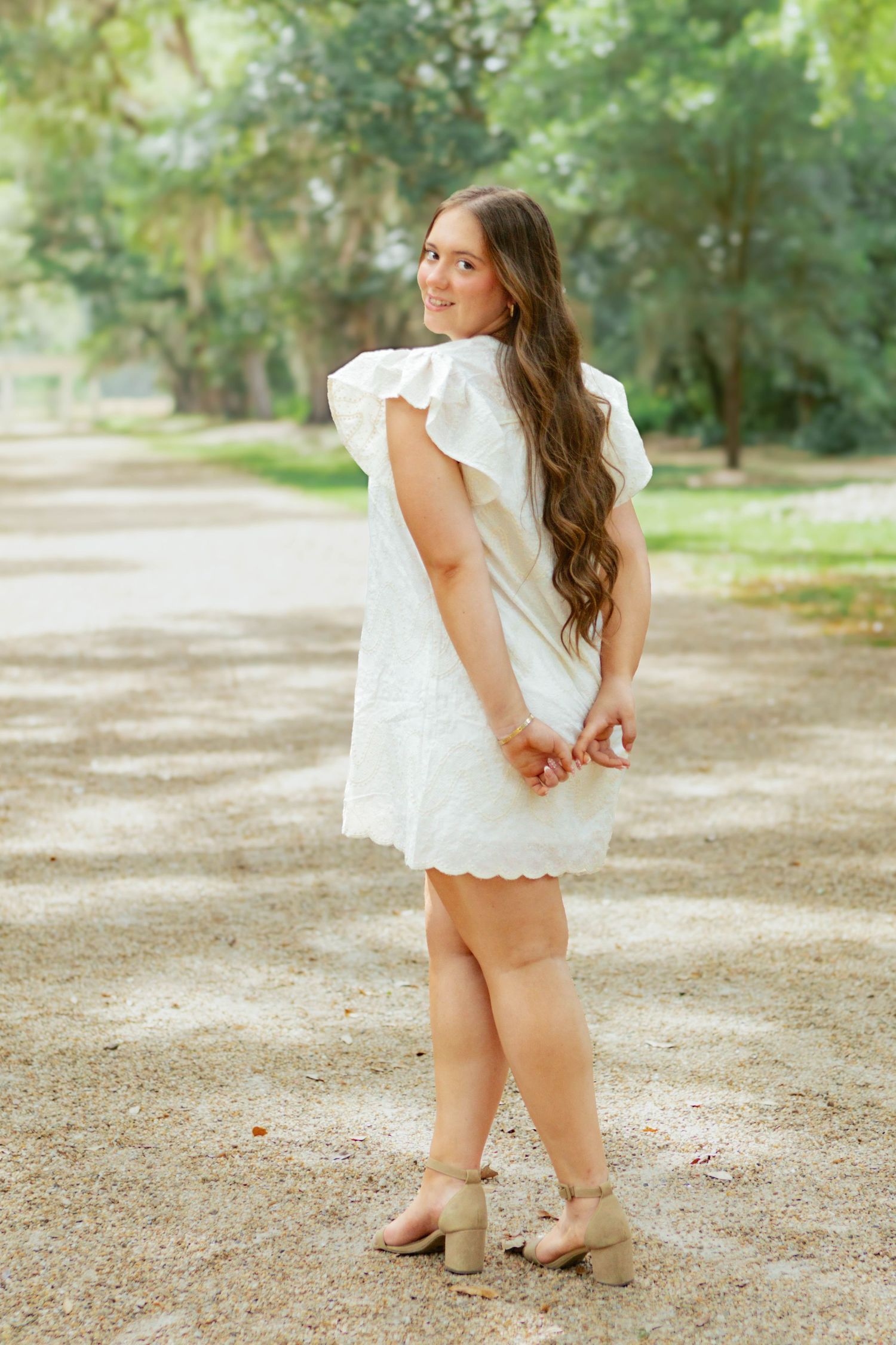 Young woman in white eyelet flutter-sleeve dress posing on tree-lined gravel path, looking over shoulder.