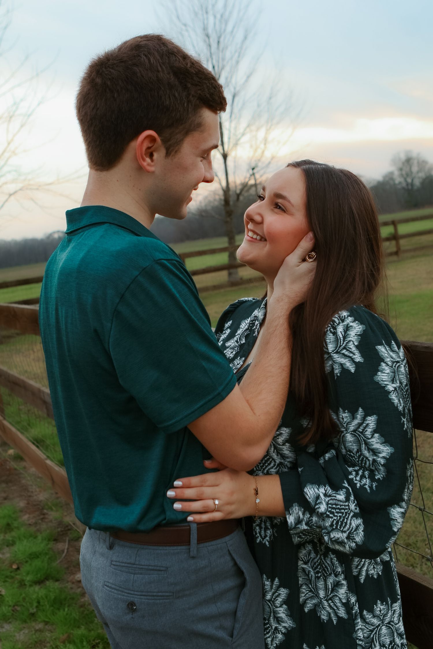 A couple shares an intimate moment together outdoors against a scenic rural backdrop with wooden fencing.