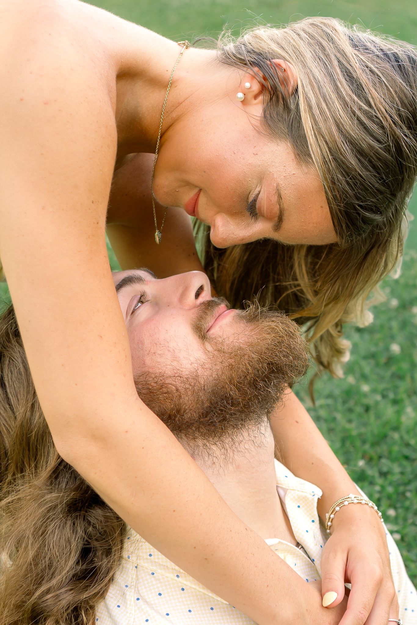 Romantic couple in a tender moment on the grass, woman leaning down toward bearded man lying below her.