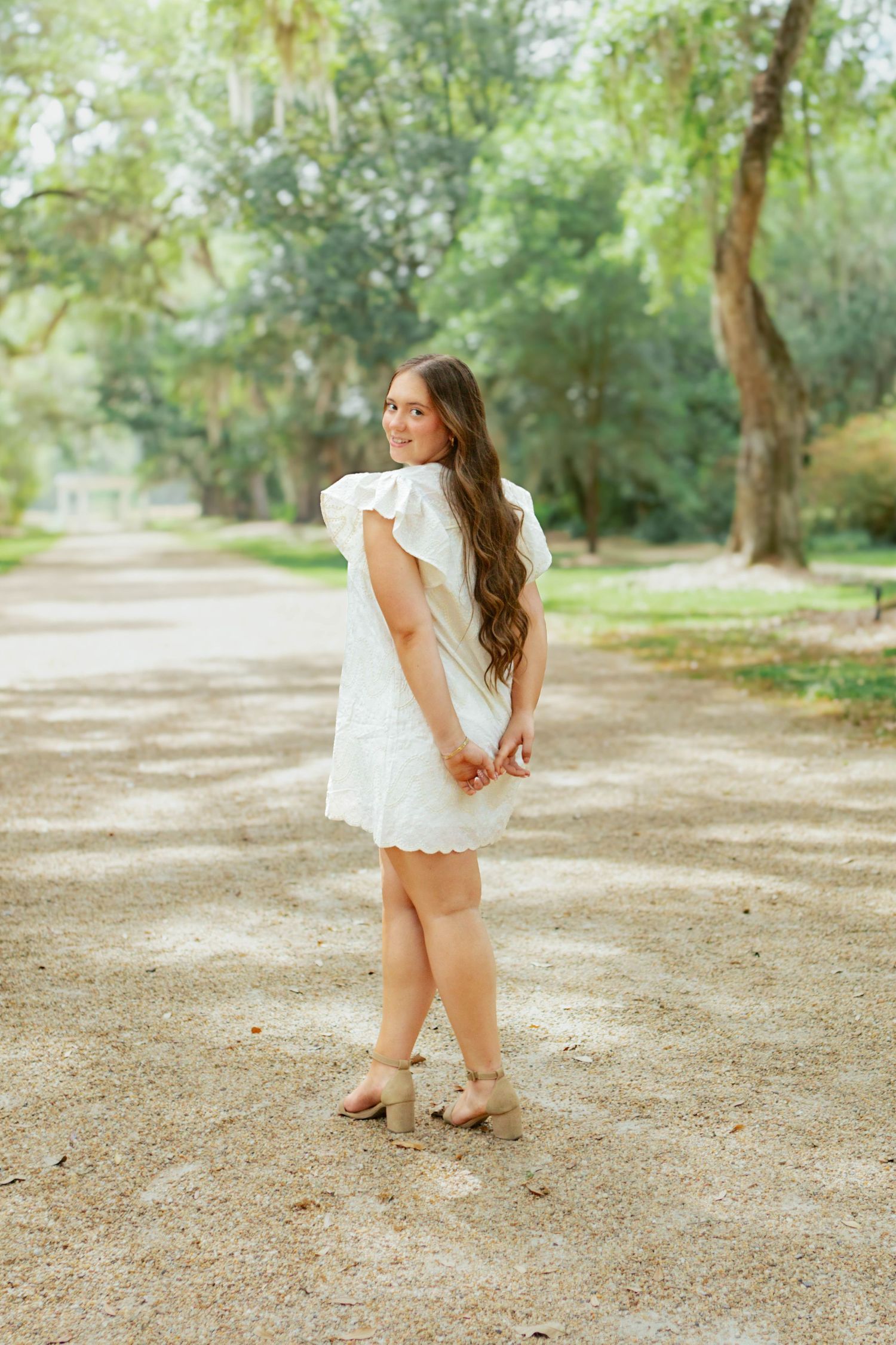 Young woman in a white eyelet flutter-sleeve dress poses on a tree-lined gravel path wearing nude heels.