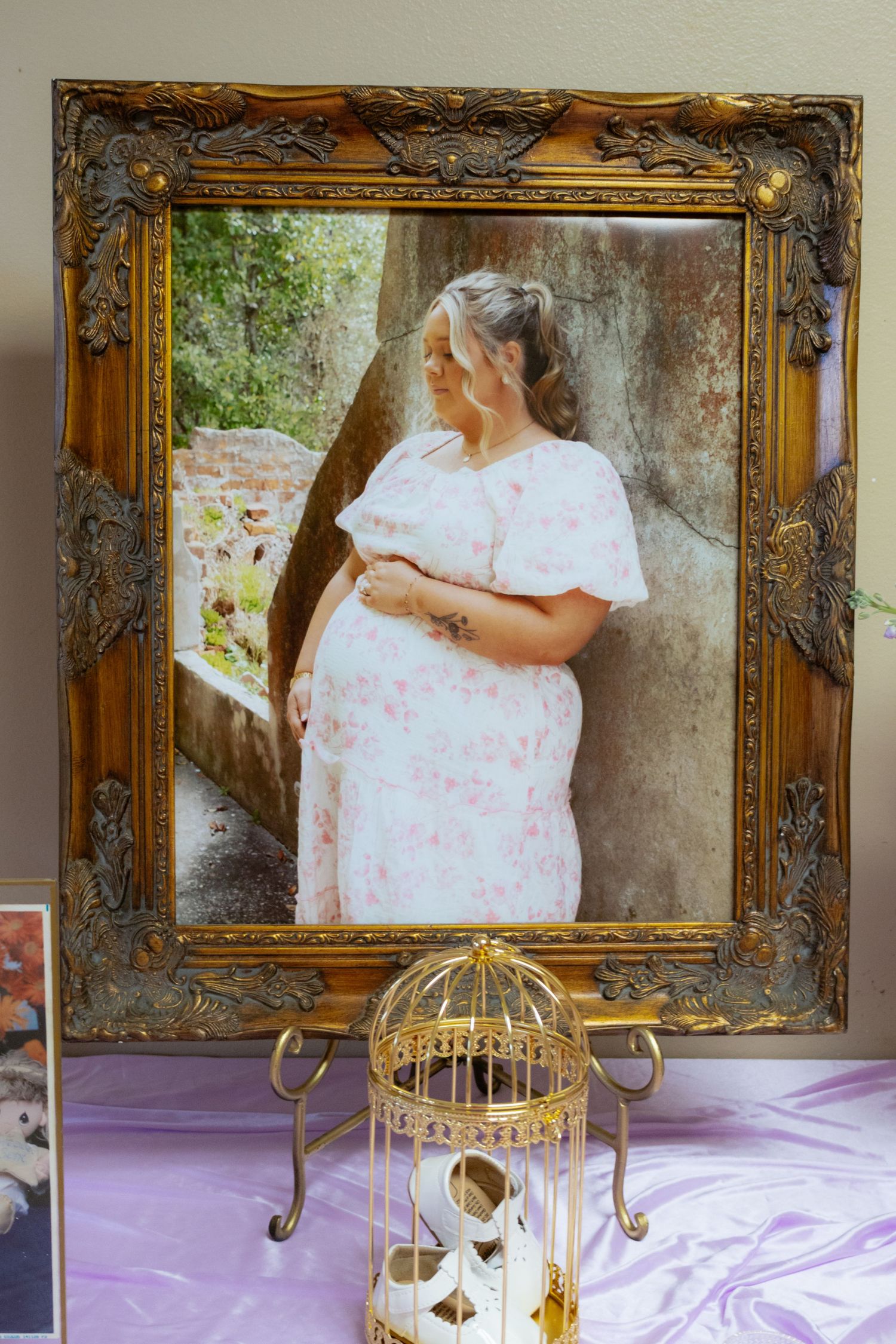 Ornate gold framed portrait of a pregnant woman in a floral dress displayed on a table with a decorative birdcage.