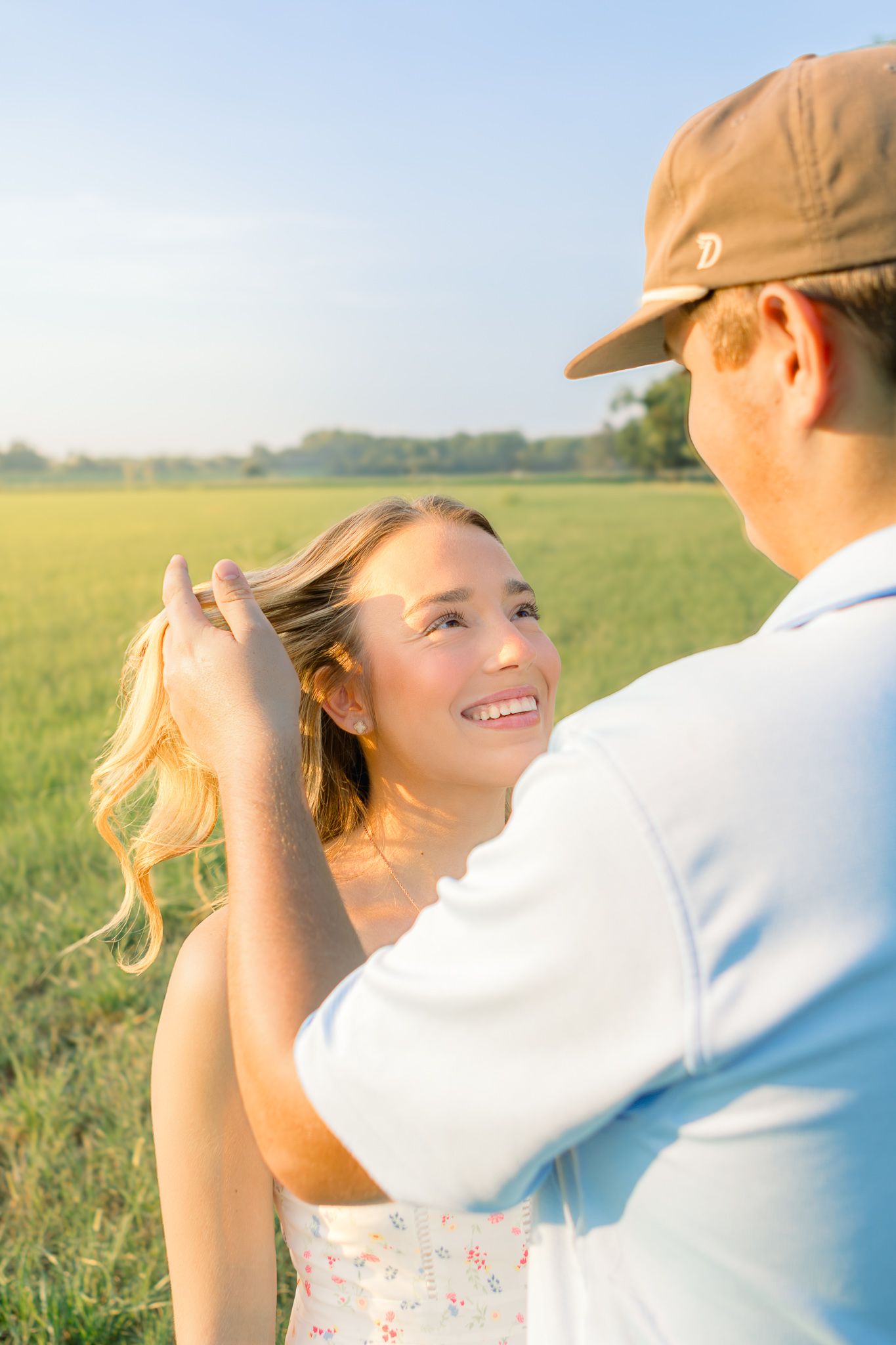 A smiling young woman looks up at a man fixing her blonde hair in a sunny green field.