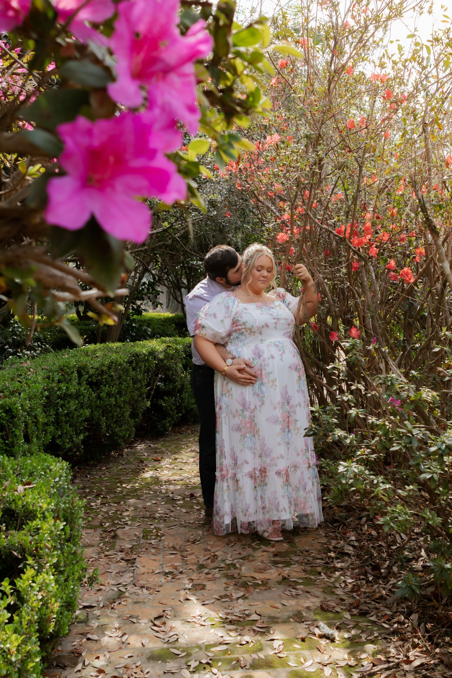 Pregnant woman in floral dress with partner embracing her in a garden surrounded by pink azalea blooms.
