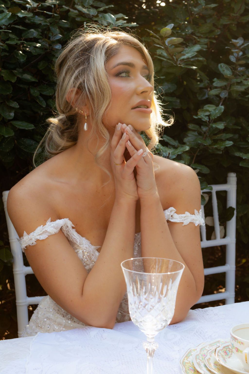 Blonde bride in an off-shoulder lace gown sitting at an outdoor table with crystal glassware in garden setting.