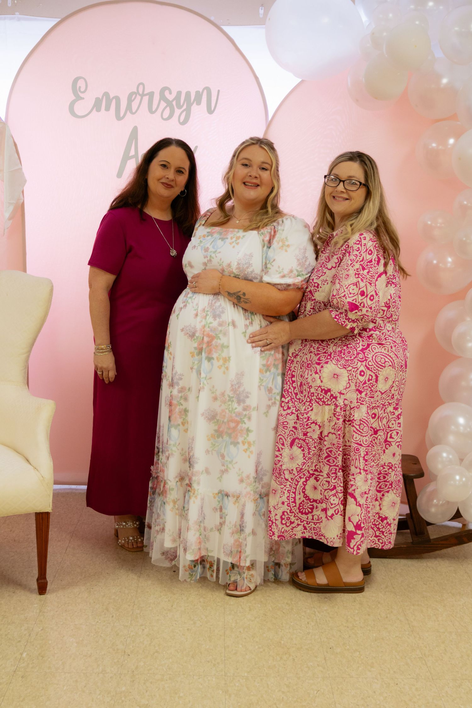 Three women posing together at Emersyn baby shower with pink balloon garland backdrop decoration.