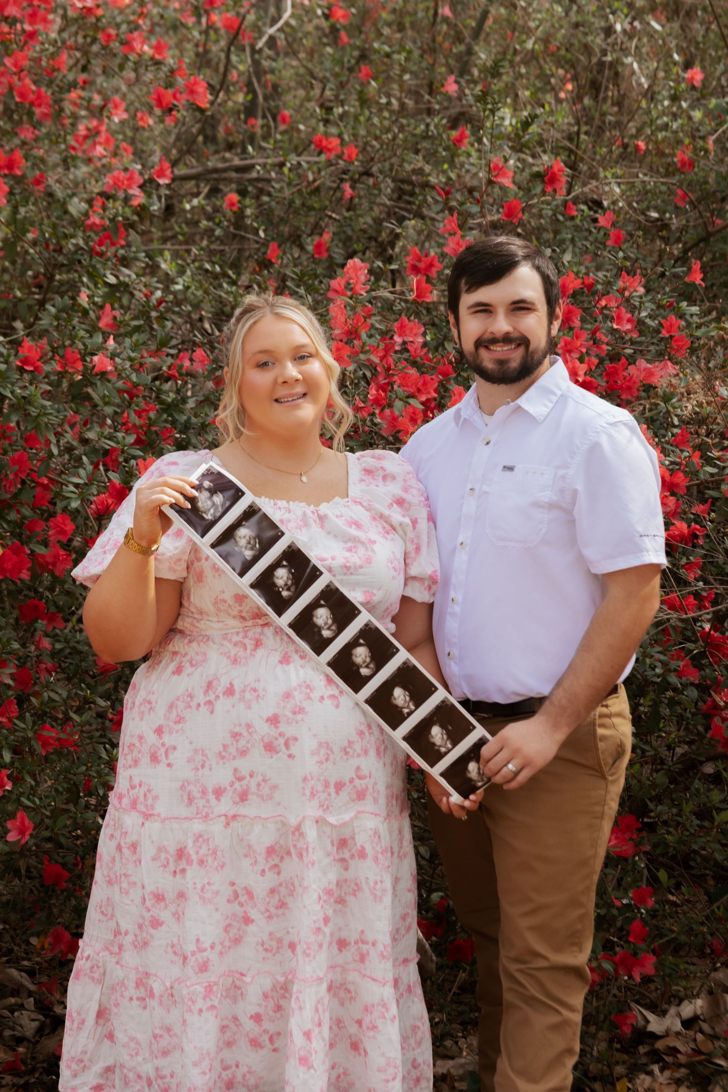 Smiling couple holding ultrasound photos in front of red flowering bushes, announcing pregnancy in a floral dress and white shirt.