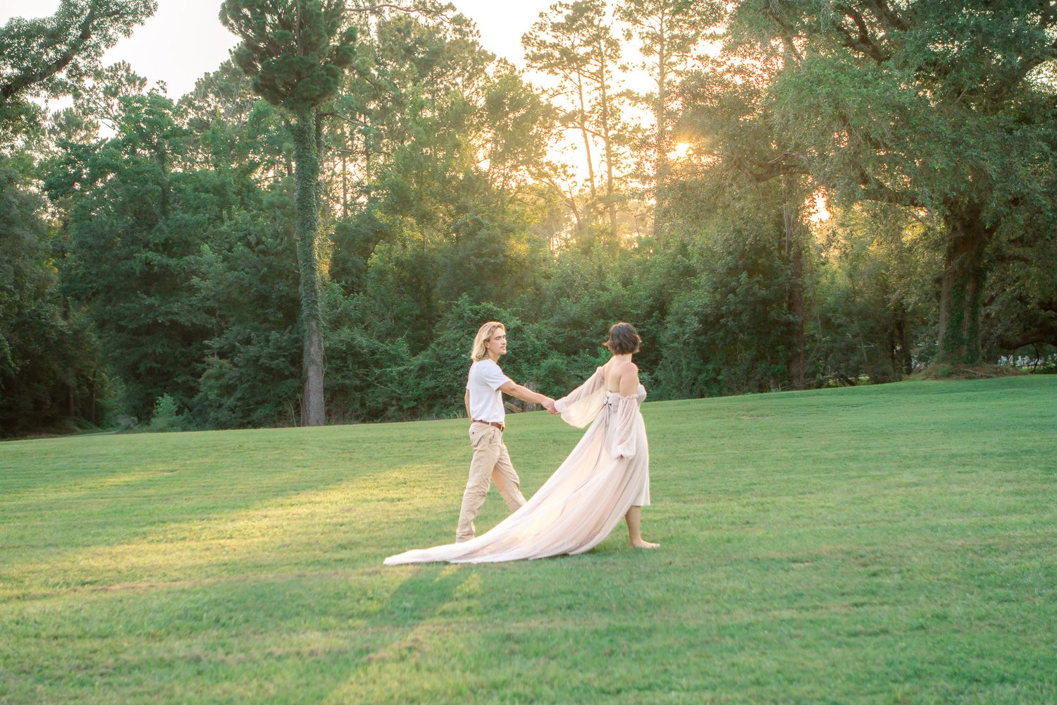 Couple holding hands on lush green lawn at sunset with tall trees and golden light in background.
