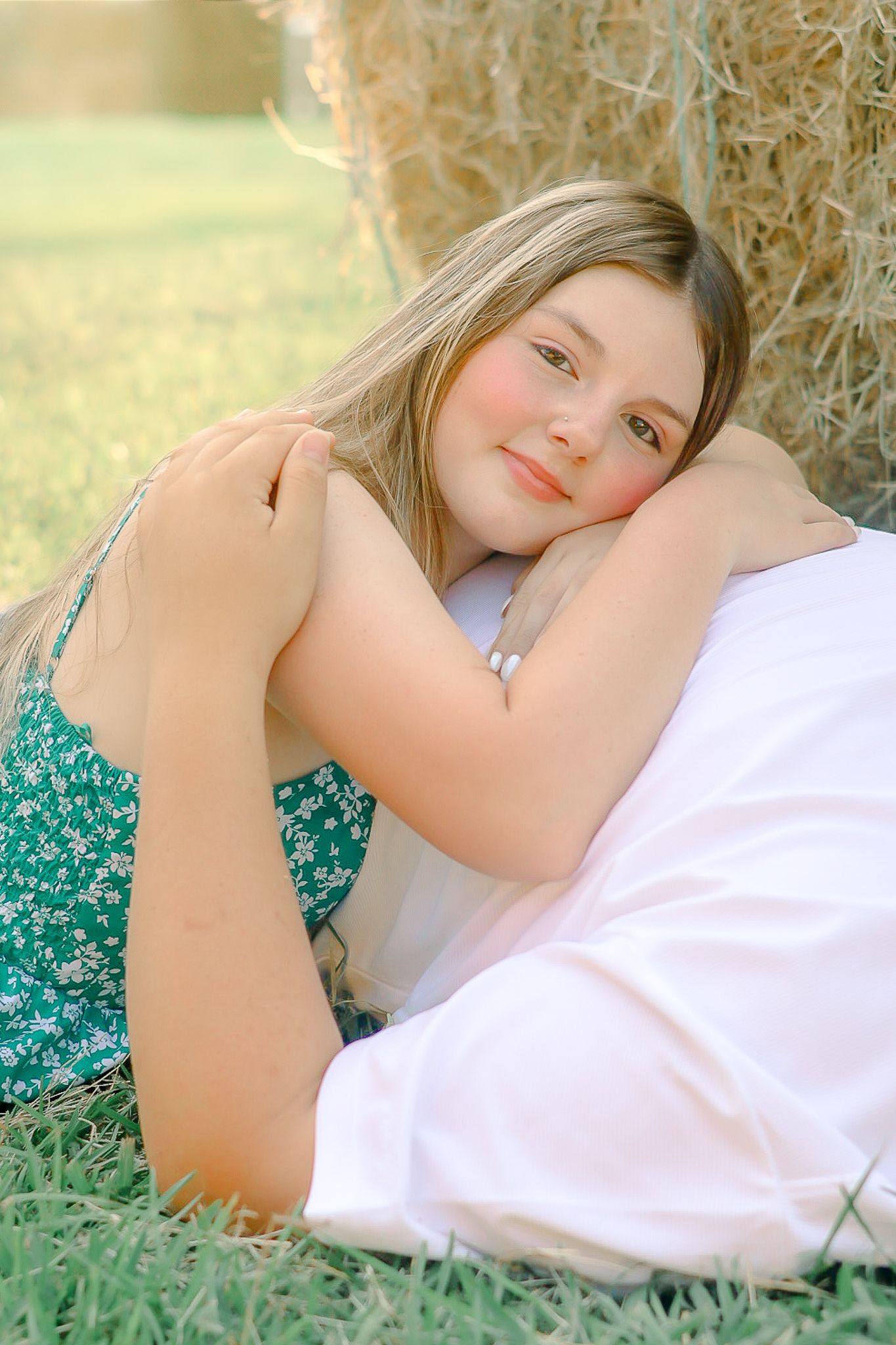 Young blonde girl in a green floral dress resting against a hay bale in a sunny field.