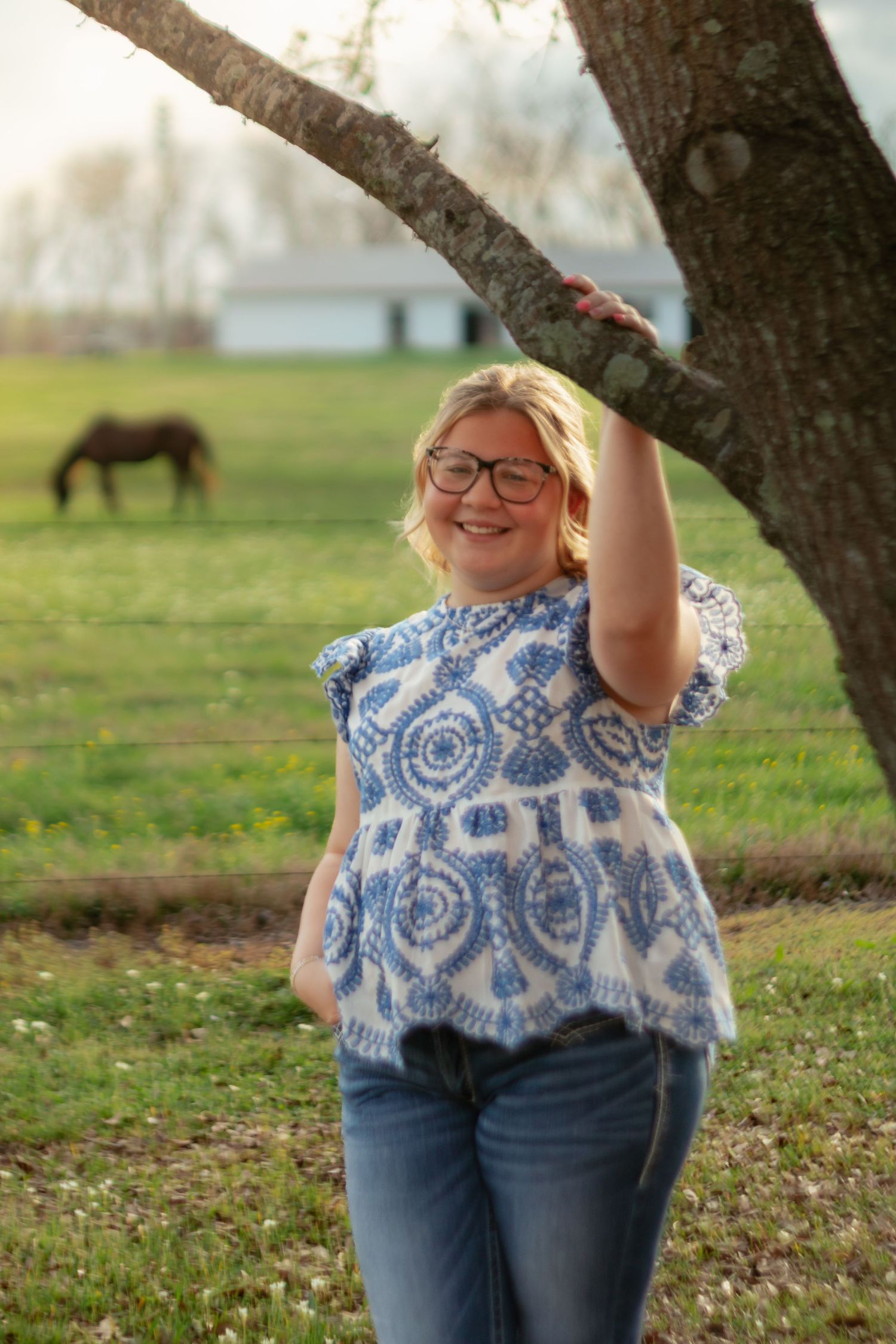 Smiling blonde woman in blue patterned top and jeans holding a tree branch in a pastoral farm setting.
