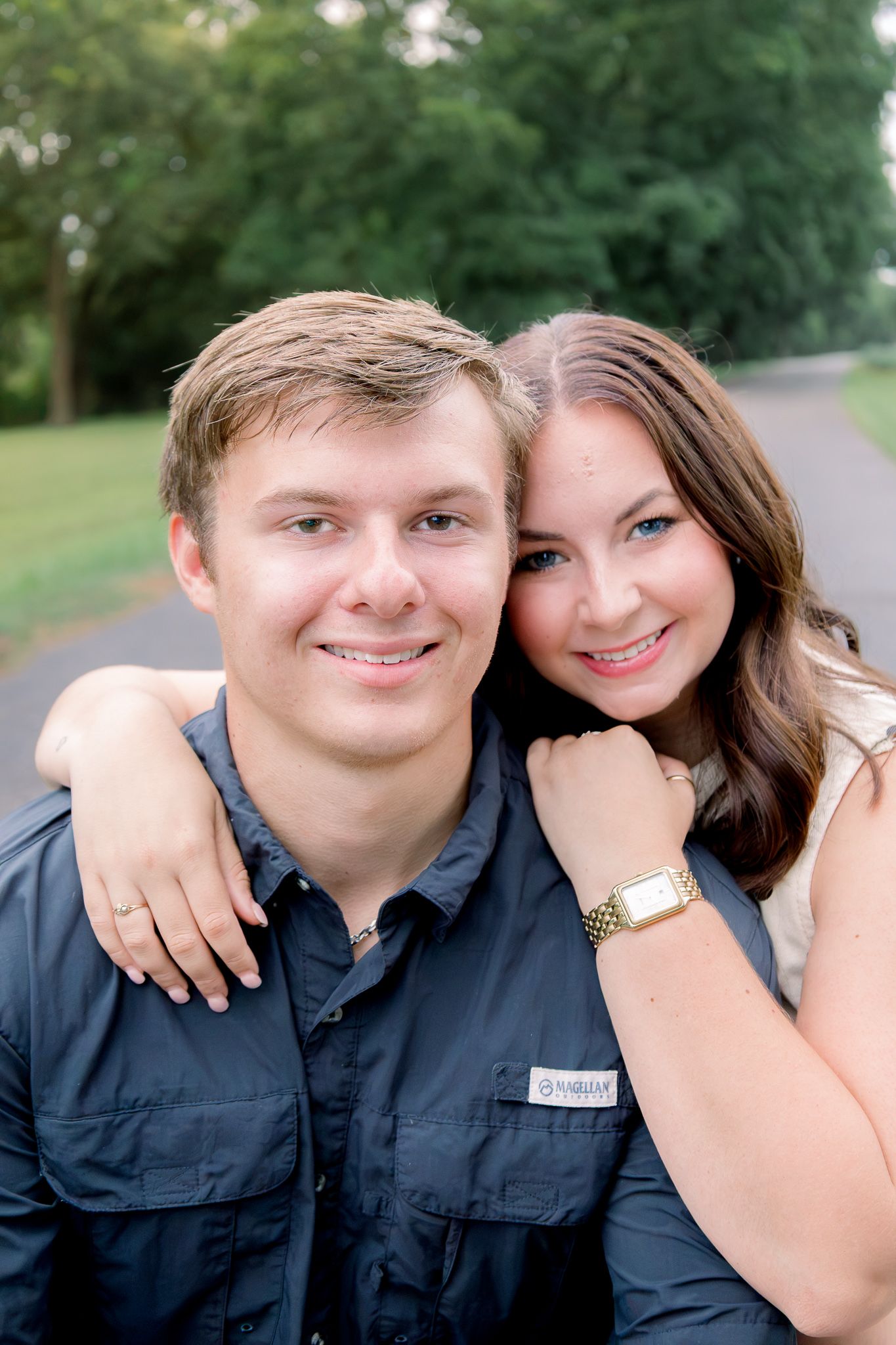 Smiling young couple posing outdoors, woman resting arm on man's shoulder, green trees in background.