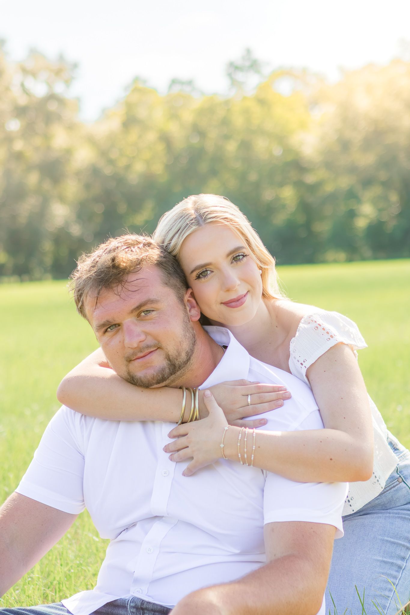 A couple sits together in a sunny green field, the woman hugging the man from behind, both smiling warmly.