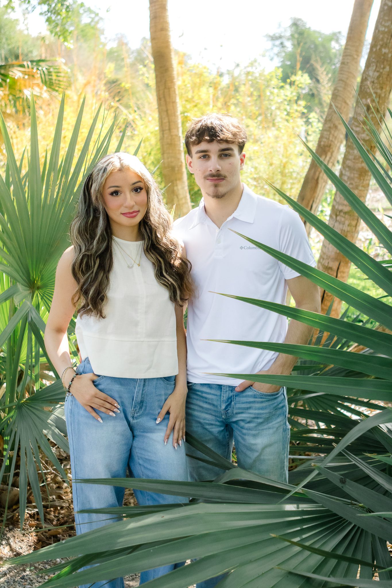 Young couple posing together among tropical palm plants in a sunny outdoor garden setting.