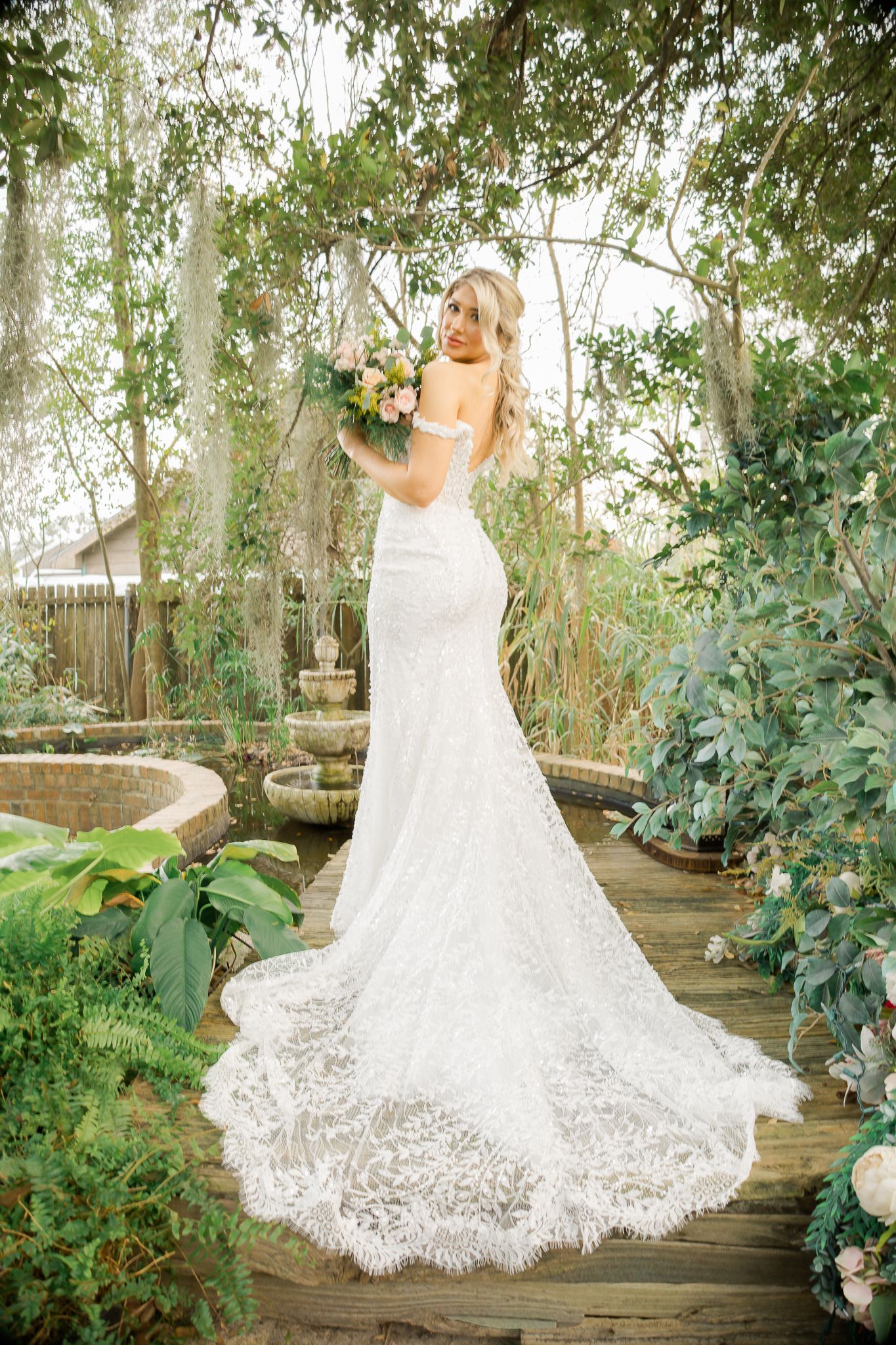 Bride in lace mermaid wedding gown with train holds bouquet in lush garden setting.
