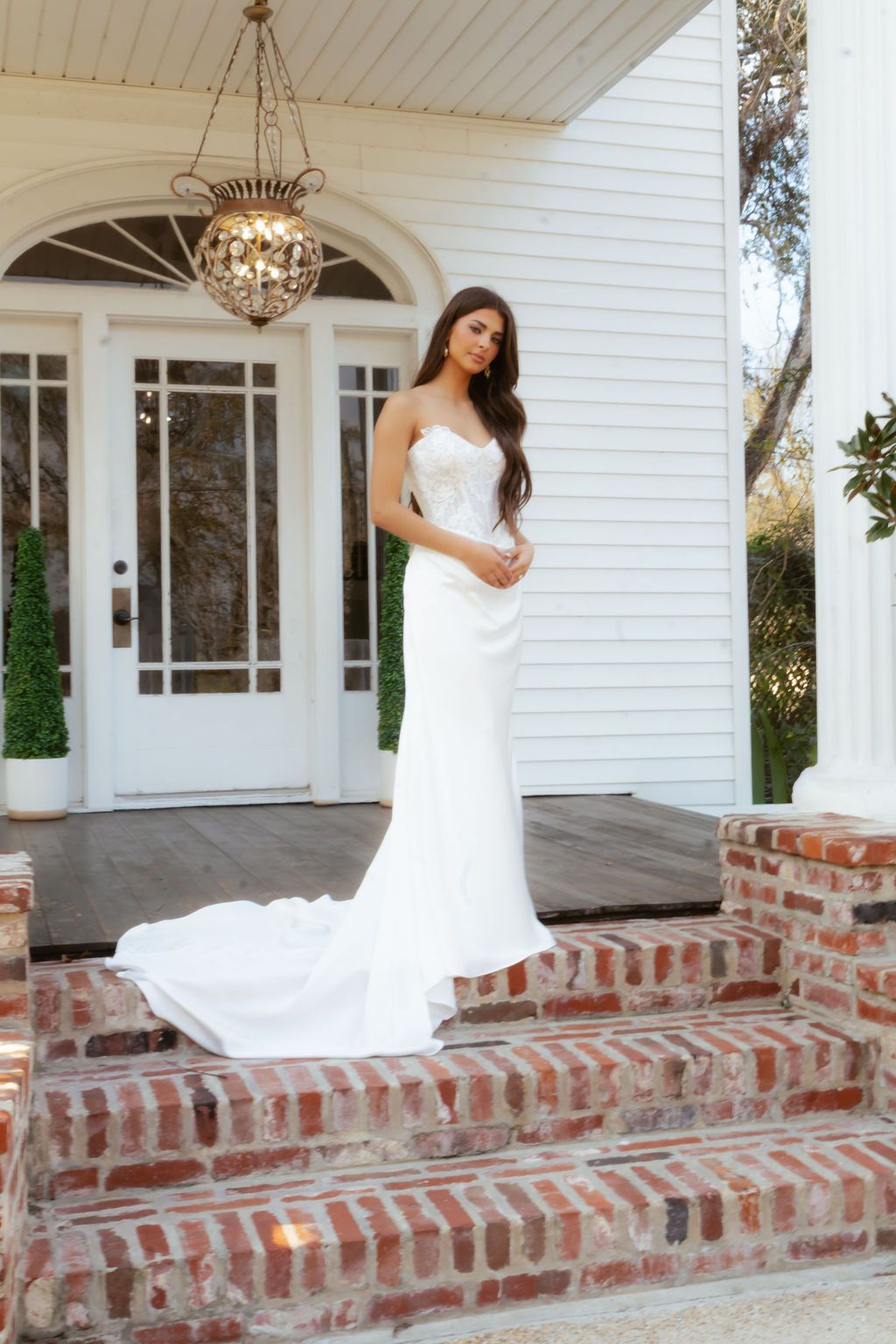 Elegant bride in a strapless white crepe gown with train standing on brick steps of a white colonial building.