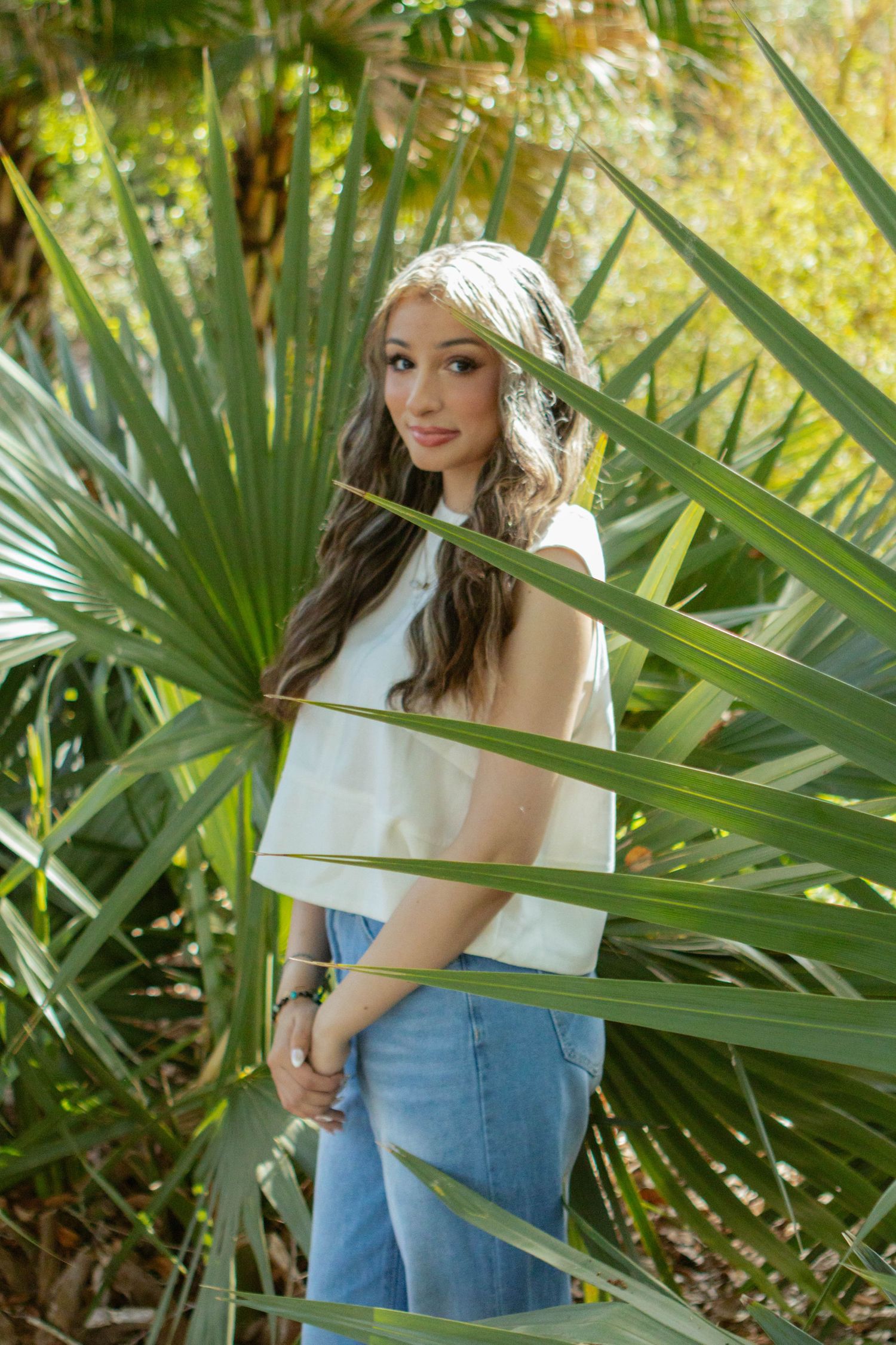 Young woman in white top and jeans posing among tropical palm fronds in outdoor garden setting.