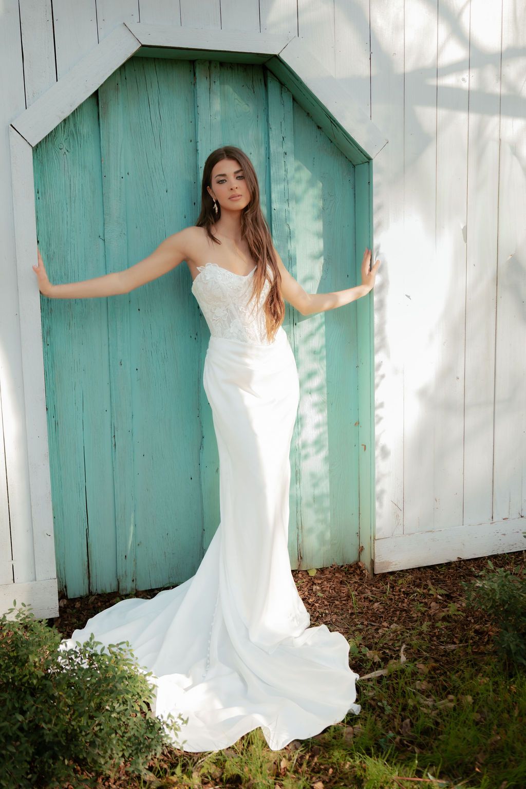 Bride in a strapless fitted white gown with train posing in a rustic turquoise wooden arched doorway outdoors.