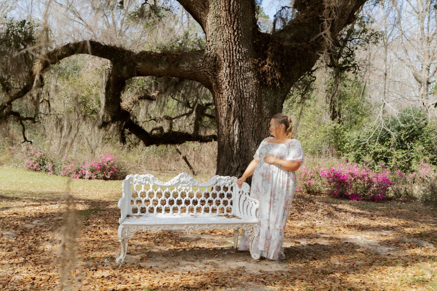 Pregnant woman in flowy dress posing by white iron bench under large oak tree with pink azaleas.