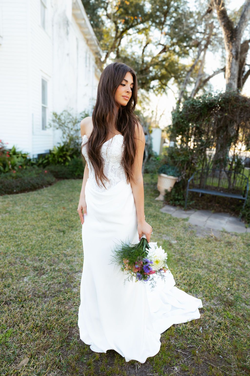 Brunette bride in strapless white mermaid gown holding wildflower bouquet in sunlit garden.