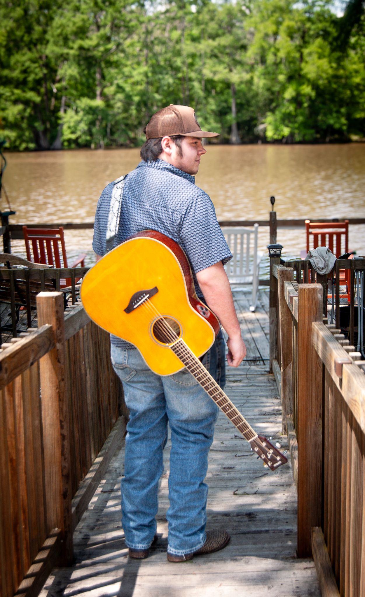 Musician with acoustic guitar on wooden dock by water wearing blue checkered shirt.