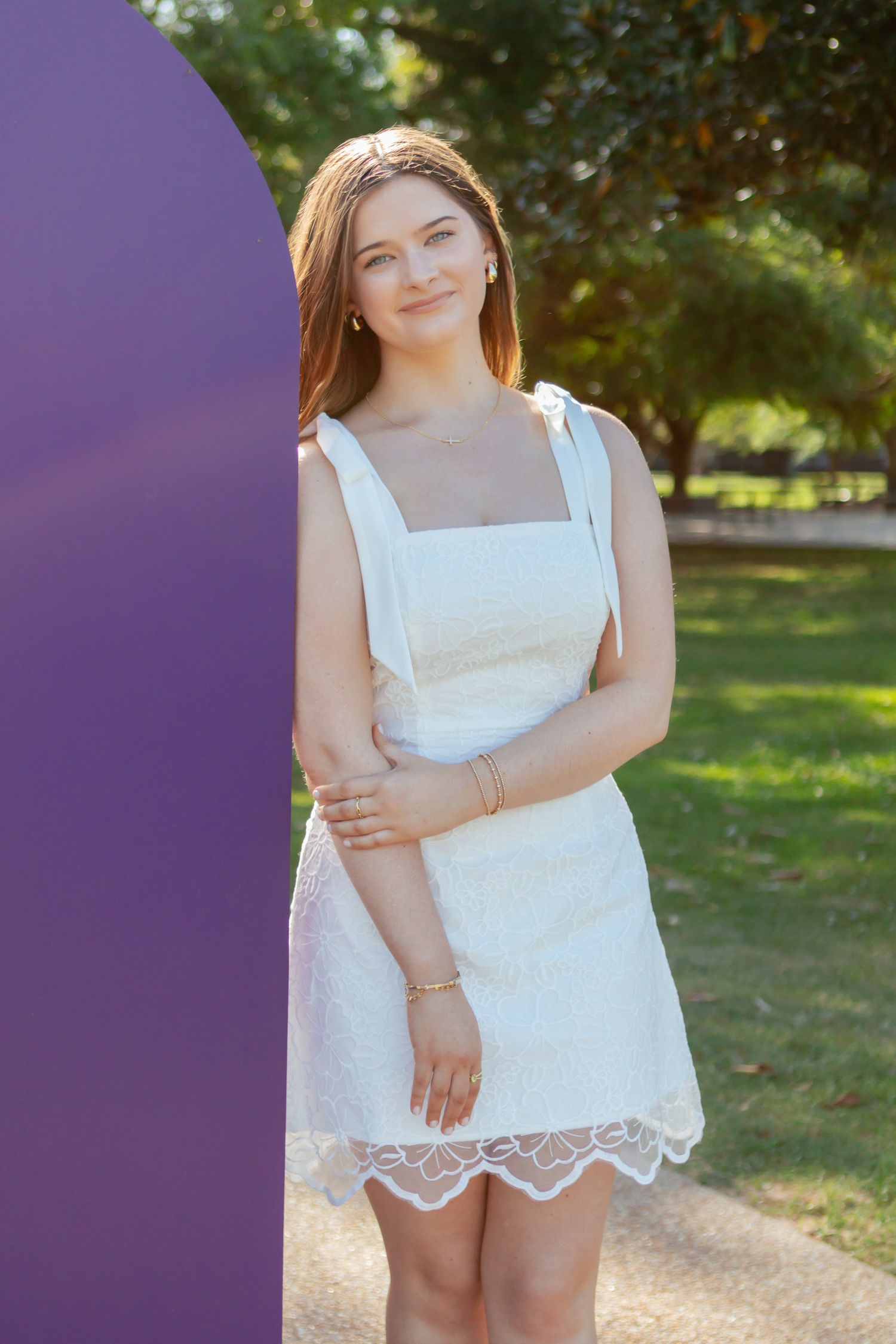 Young woman in white lace sundress posing beside purple decorative structure in sunny park setting.