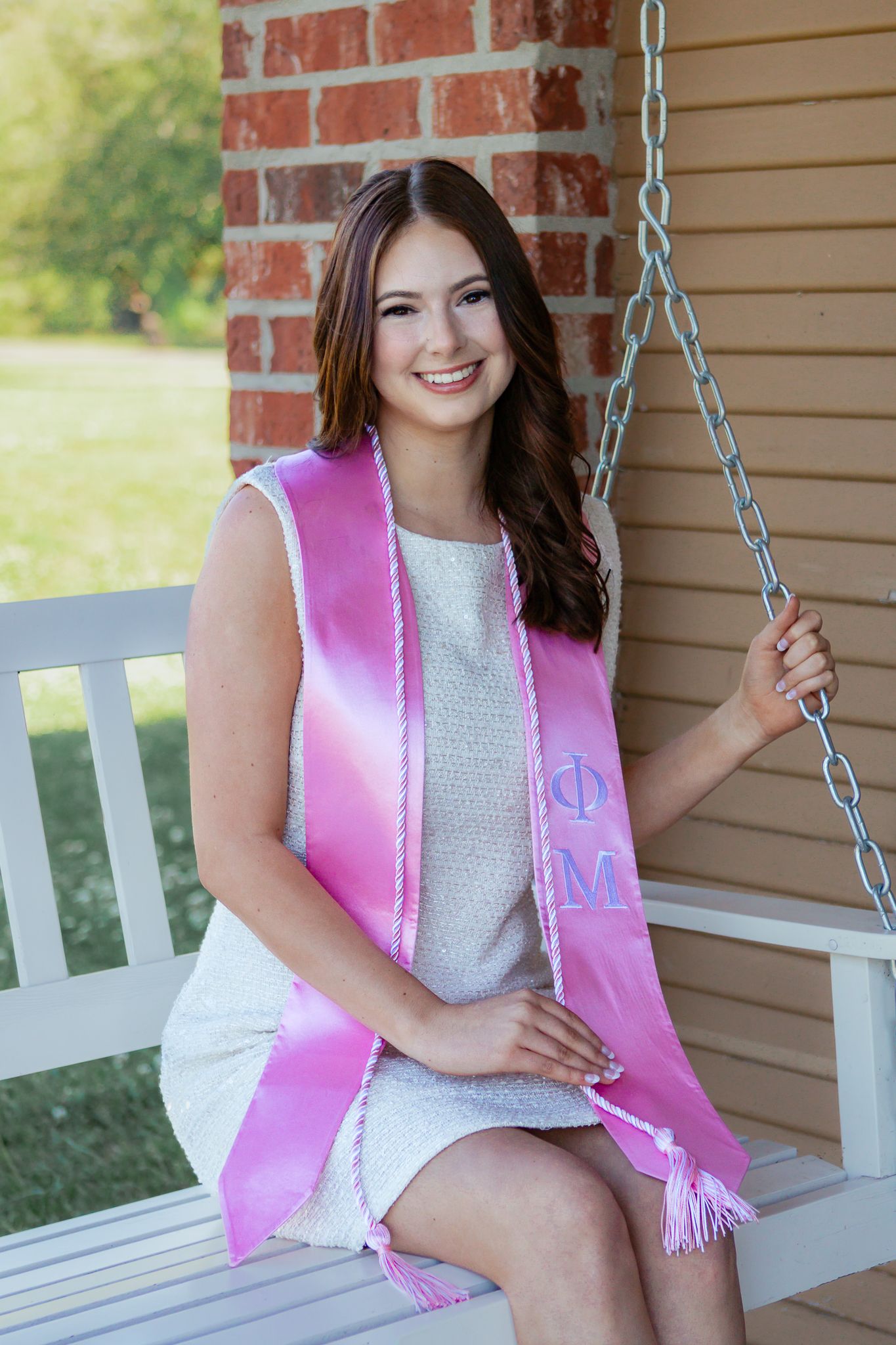 Smiling young woman wearing a pink sorority graduation sash sitting on a porch swing outdoors.
