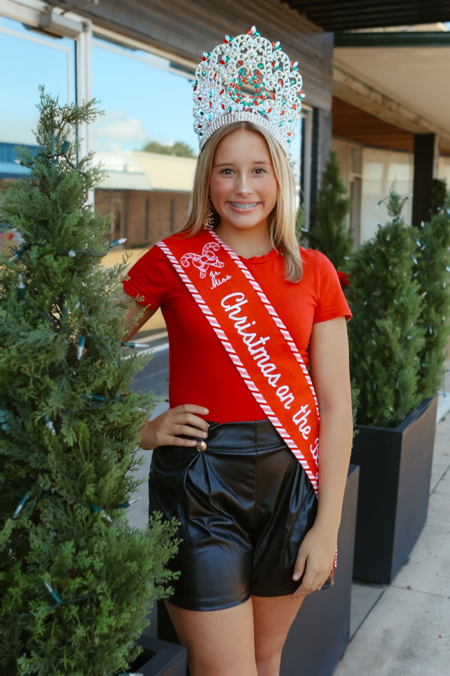Person in red shirt and black shorts poses near outdoor greenery with crown and sash.